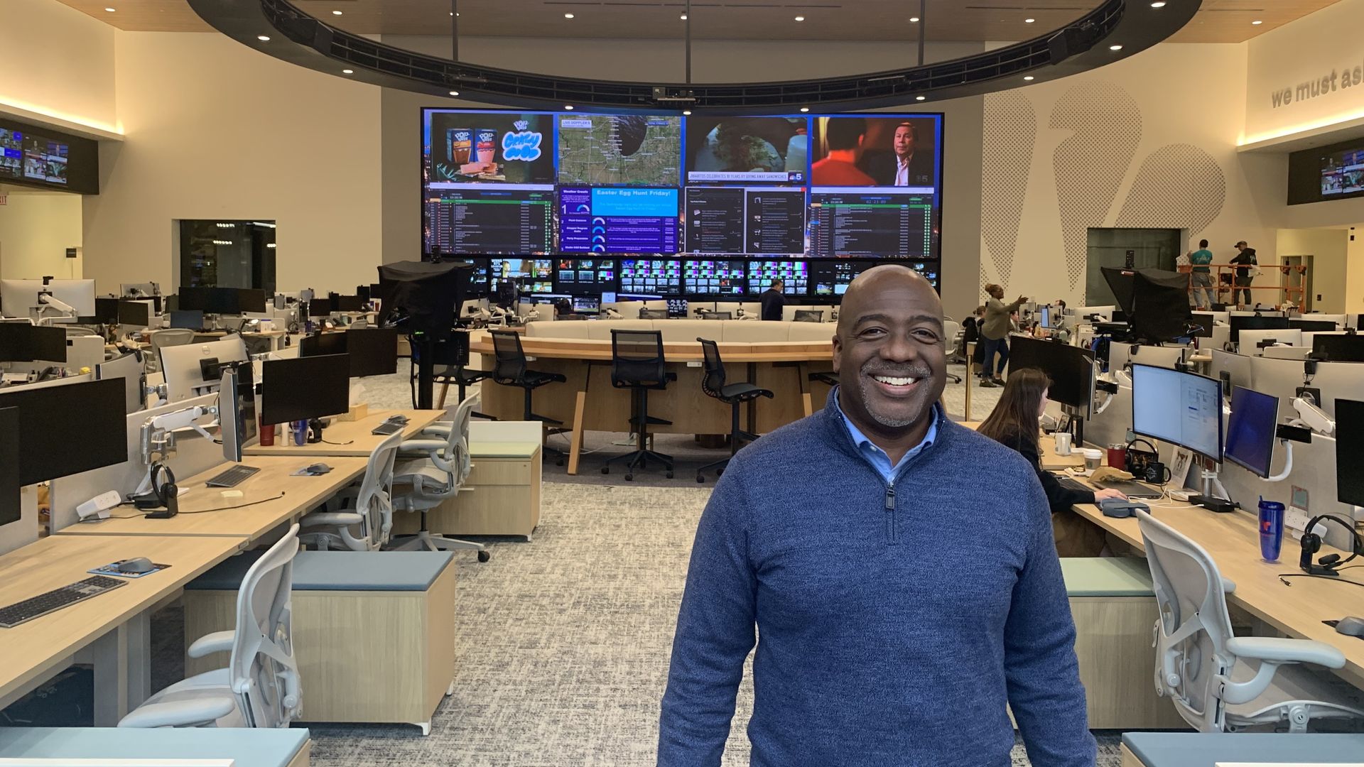 Smiling man in a blue sweater stands in a busy broadcast newsroom with rows of desks and computer monitors. A circular ticker and a large multi-screen wall loom over the space.
