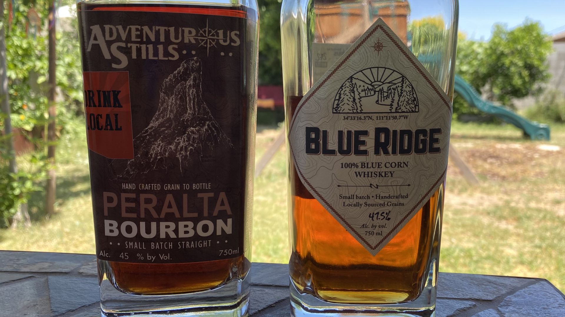 Two bottles of liquor on a table with grass and trees in the background.