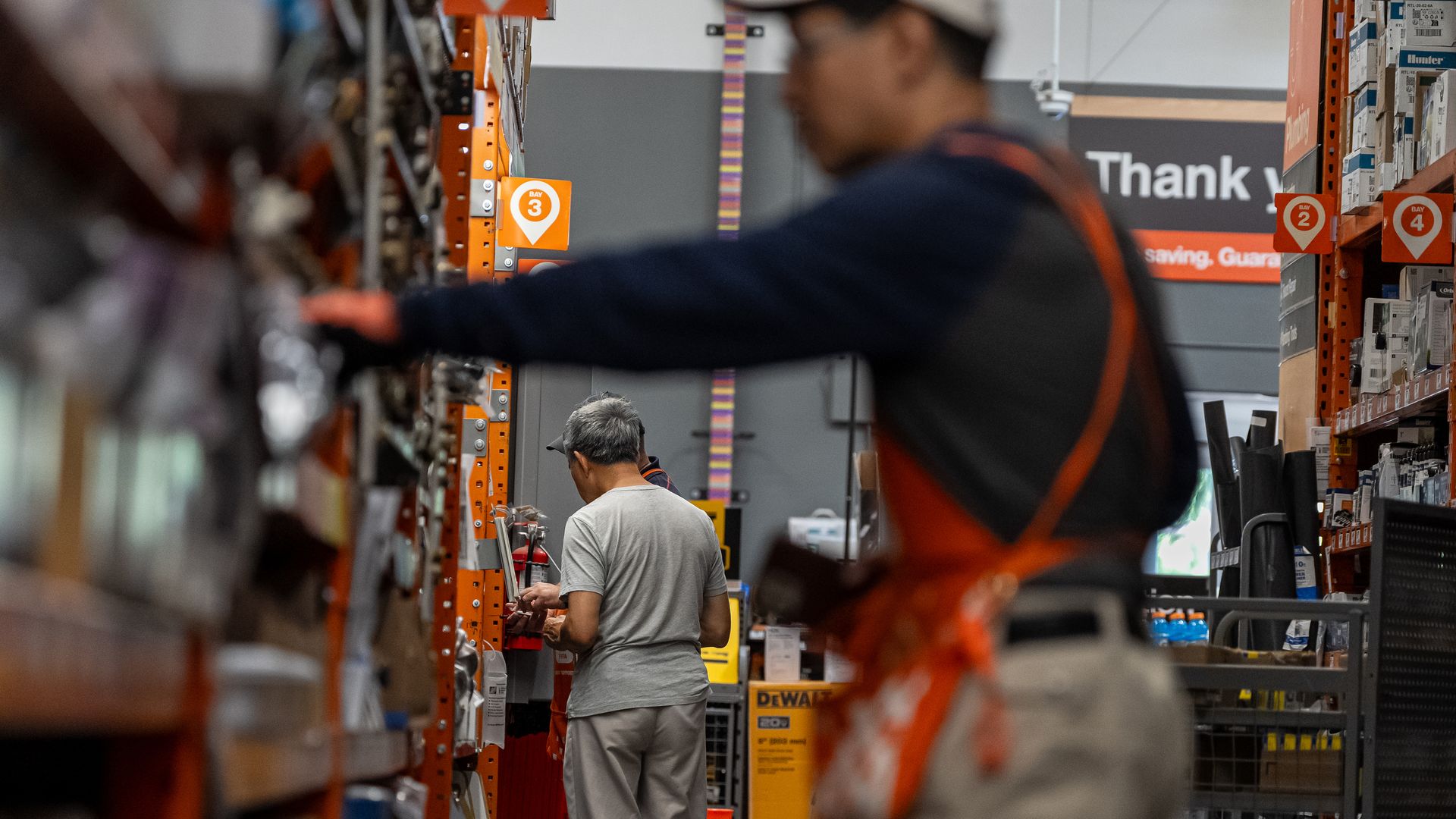 Warehouse aisle with orange shelving, a man in gray shirt facing away, a blurred foreground worker, stacked boxes, and a "Thank you" sign with bay labels 2 and 3.