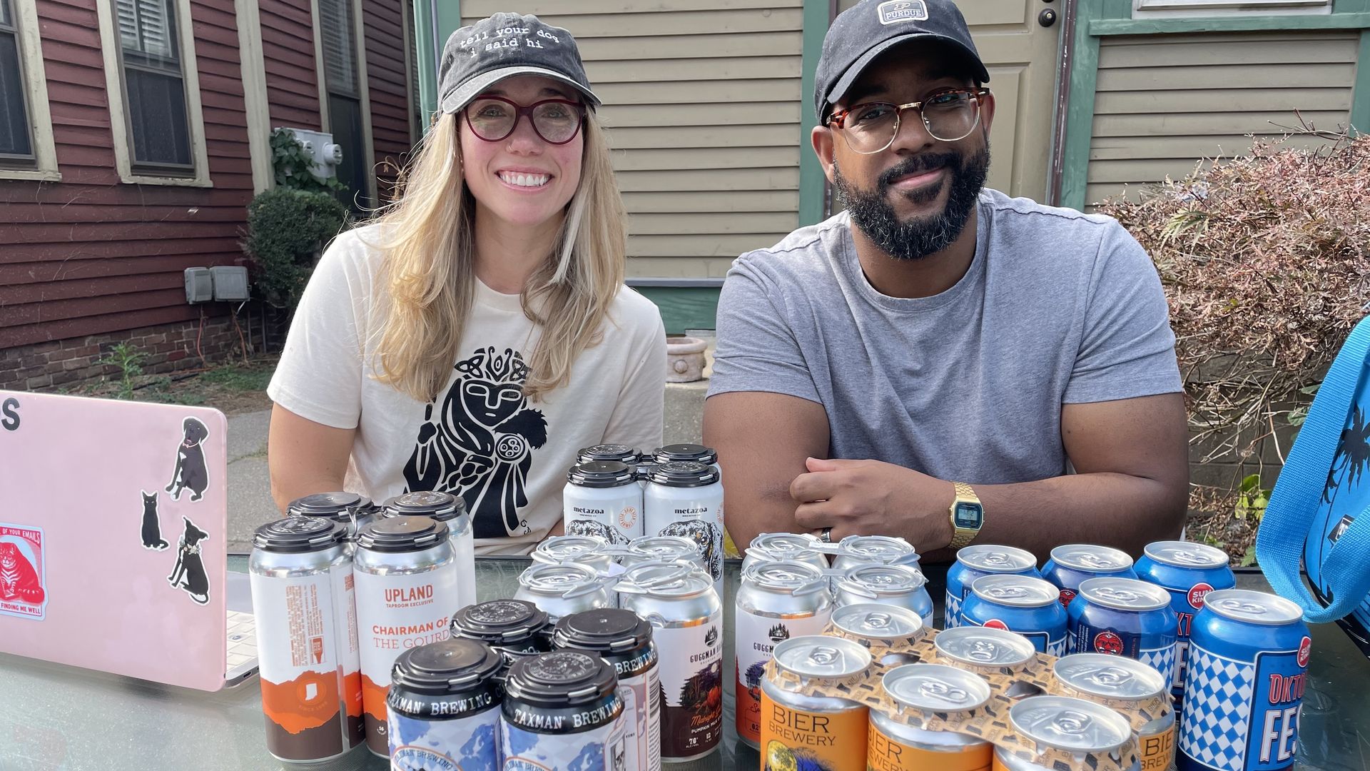 Two people wearing glasses and caps sit at a table with various craft beer cans, including labels like "BIER Brewery Pumpkin Ale" and "Expat Oktoberfest." A pink laptop with animal stickers is visible.