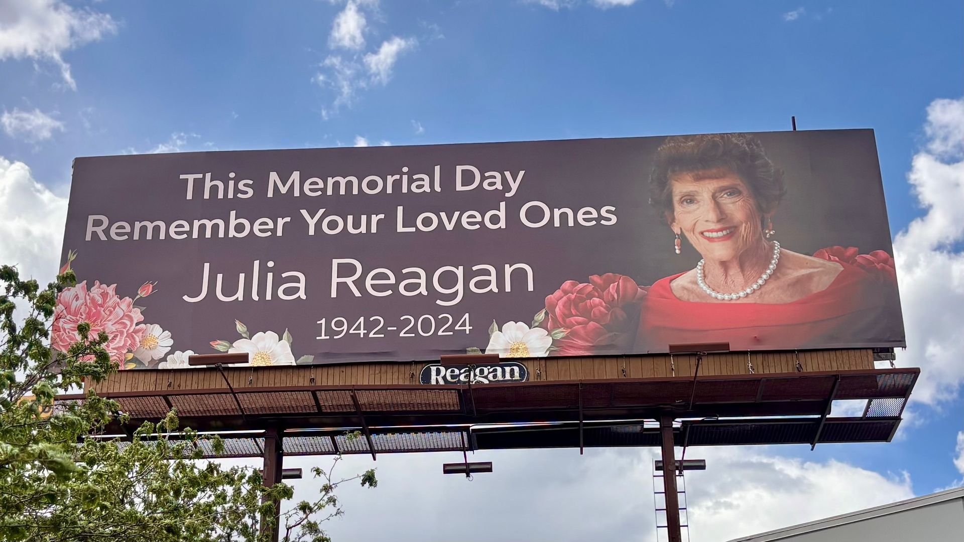Billboard memorial for Julia Reagan (1942-2024). Smiling elderly woman in a red dress with pearls on the right; text reads "This Memorial Day Remember Your Loved Ones" on a purple background.