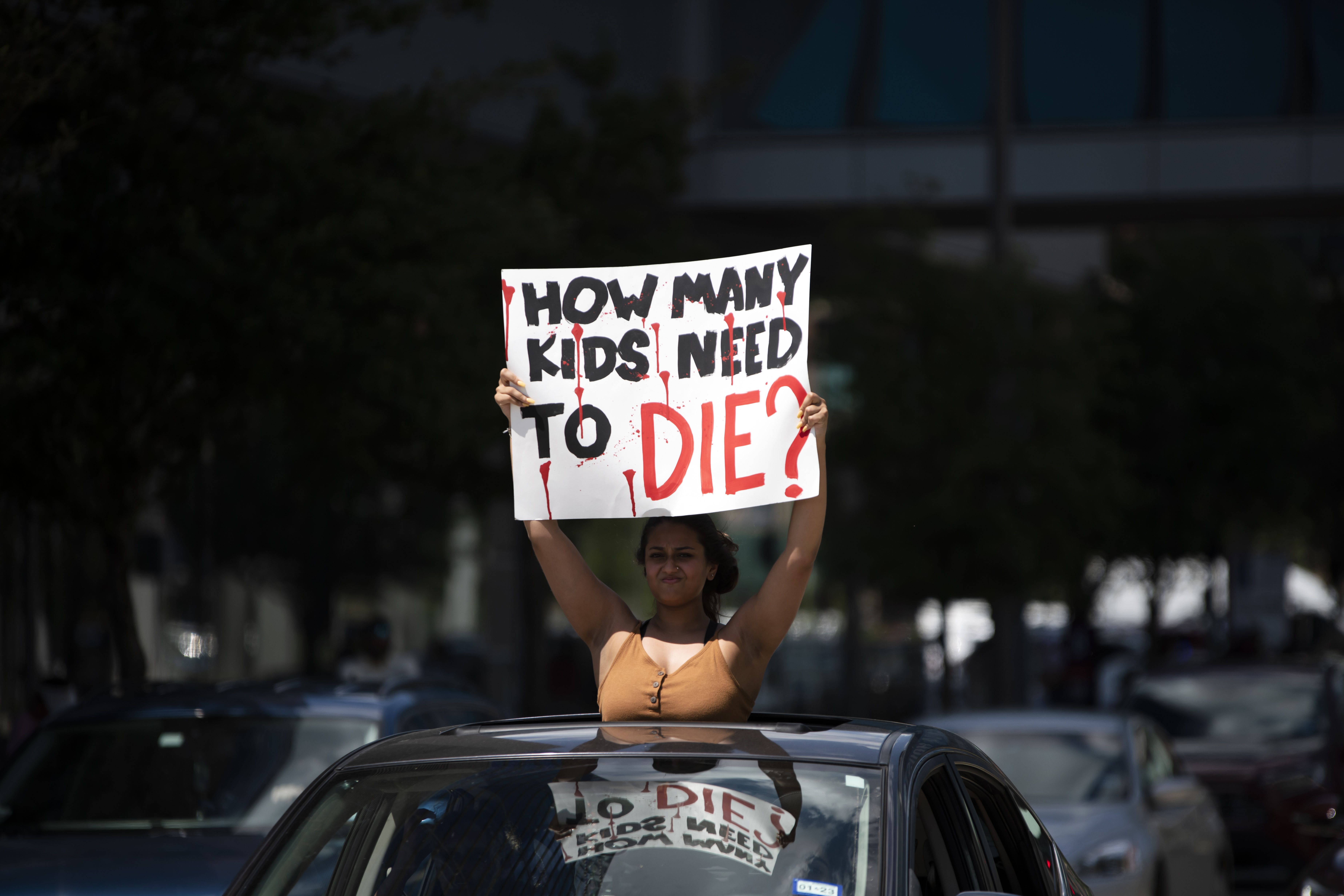 Photo of a person standing in a convertible car with a sign that says "How many kids need to die?"