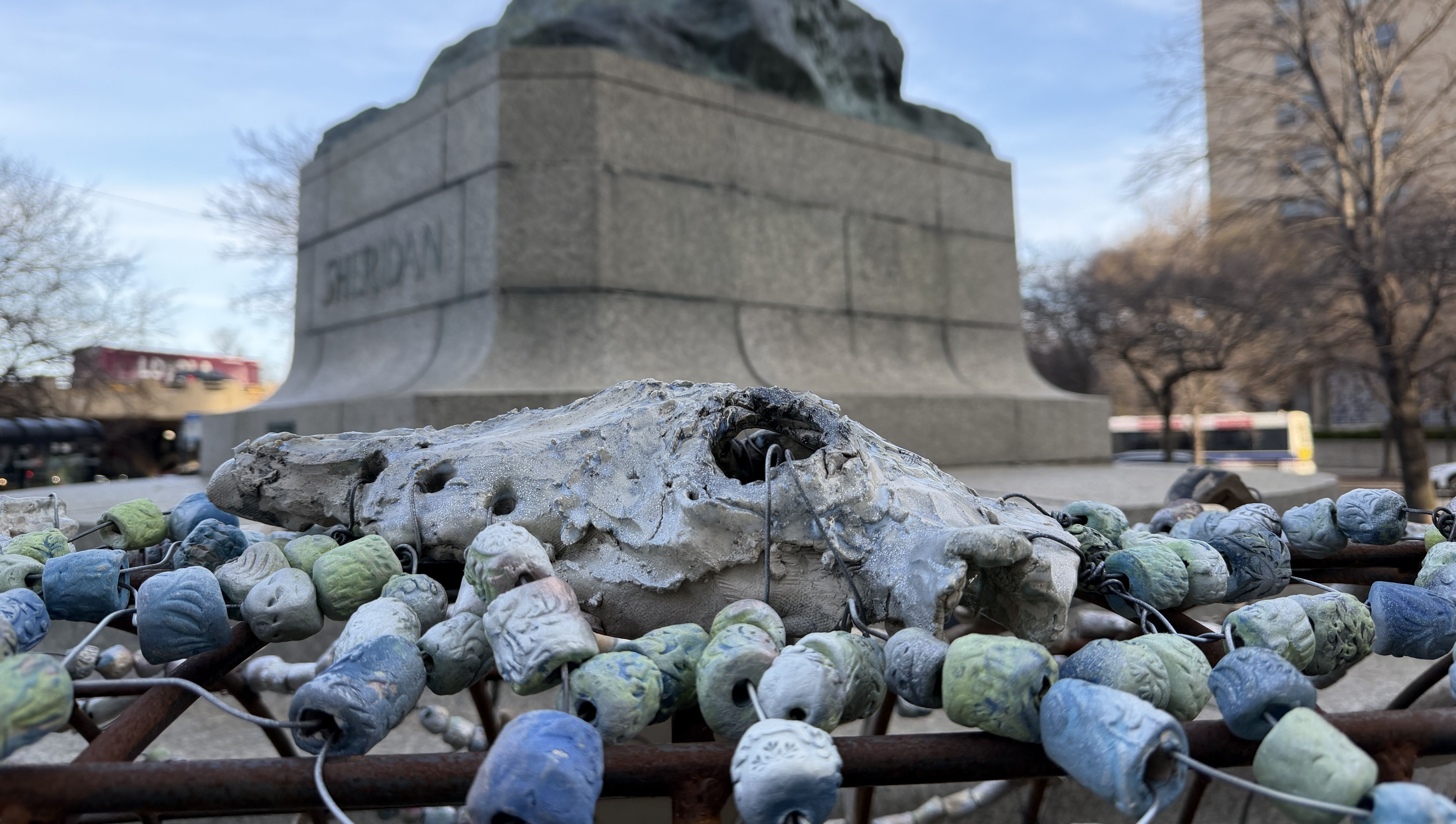Rusty railing in a park is wrapped with blue, green, and white ceramic beads around a damaged stone sculpture; a large monument sits in the background, with leafless trees and a building nearby.