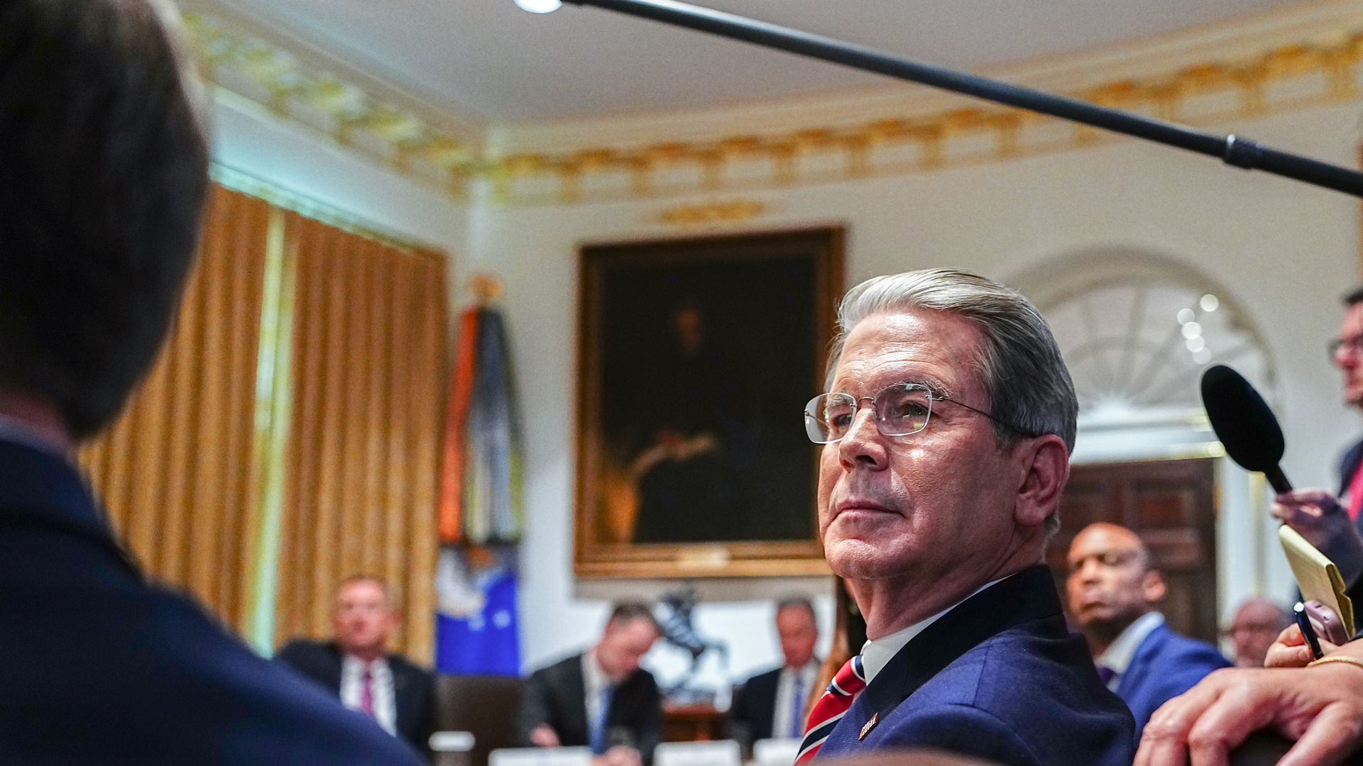Man in a dark blue suit with red striped tie and glasses is seated in a formal room with gold-trimmed curtains, a portrait on the wall, and a boom mic overhead.
