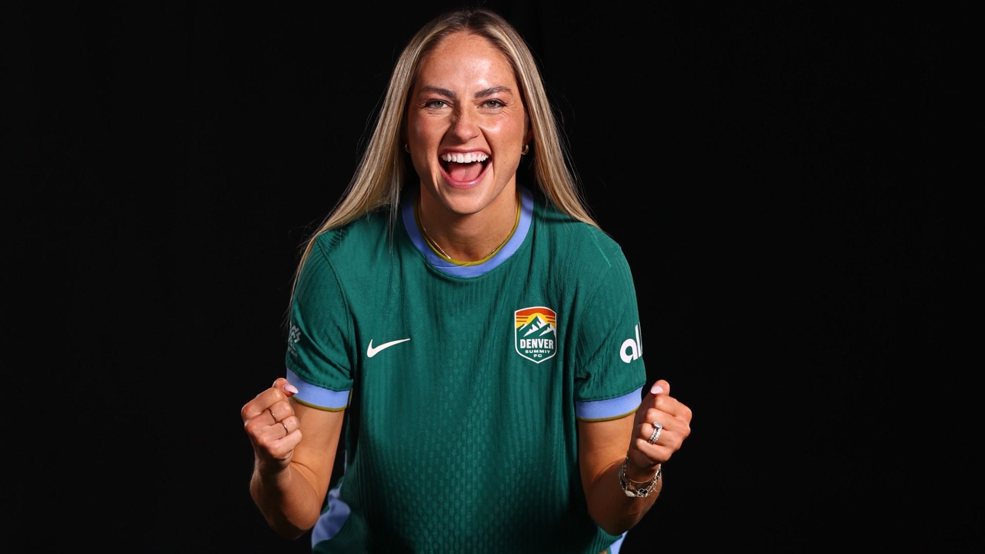 Smiling woman in a green Denver Summit FC jersey clenches fists in excitement against a black background.