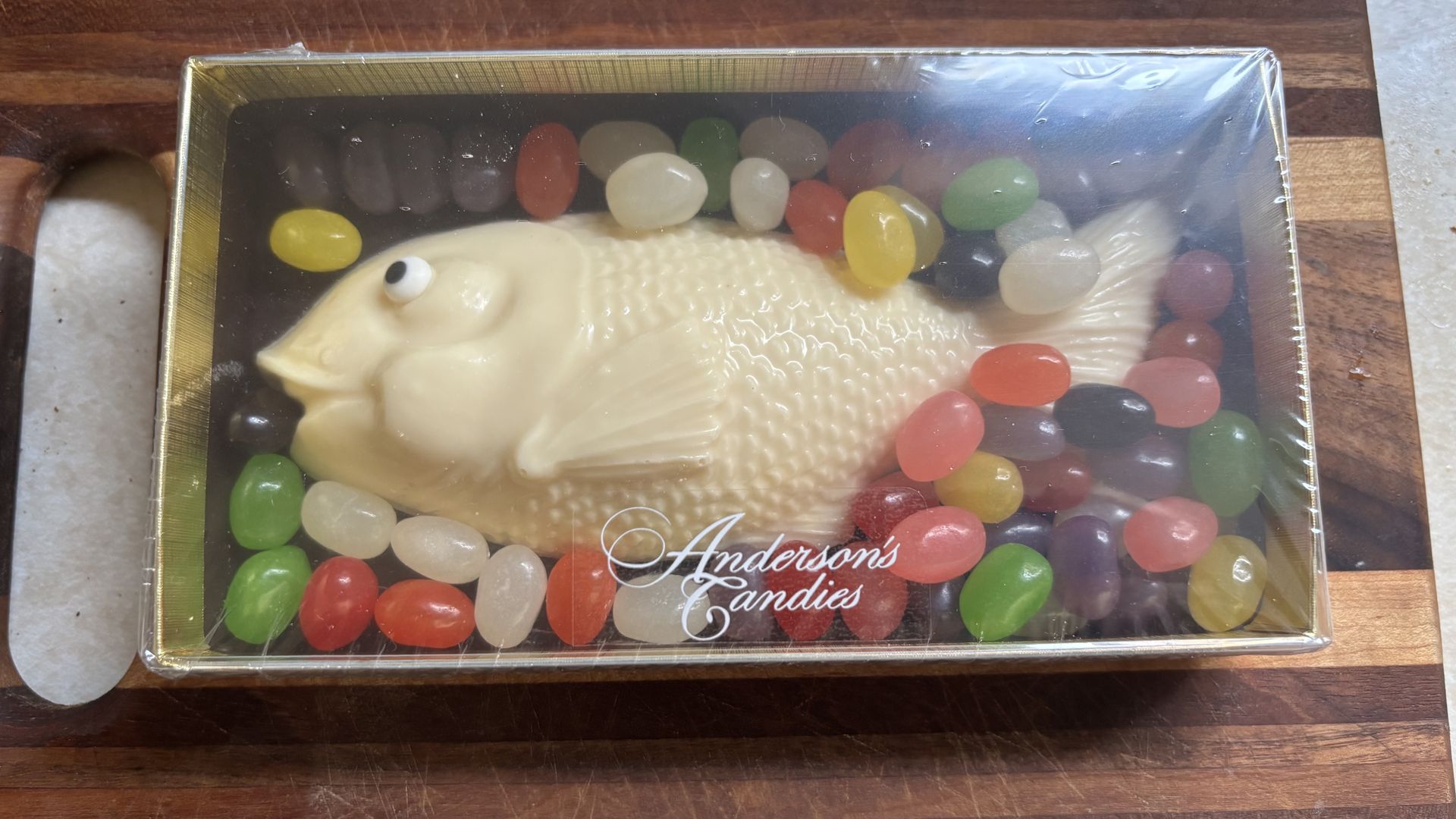 A fish-shaped white chocolate candy sits inside a clear tin box surrounded by colorful jelly beans; the box is labeled "Anderson's Candies".