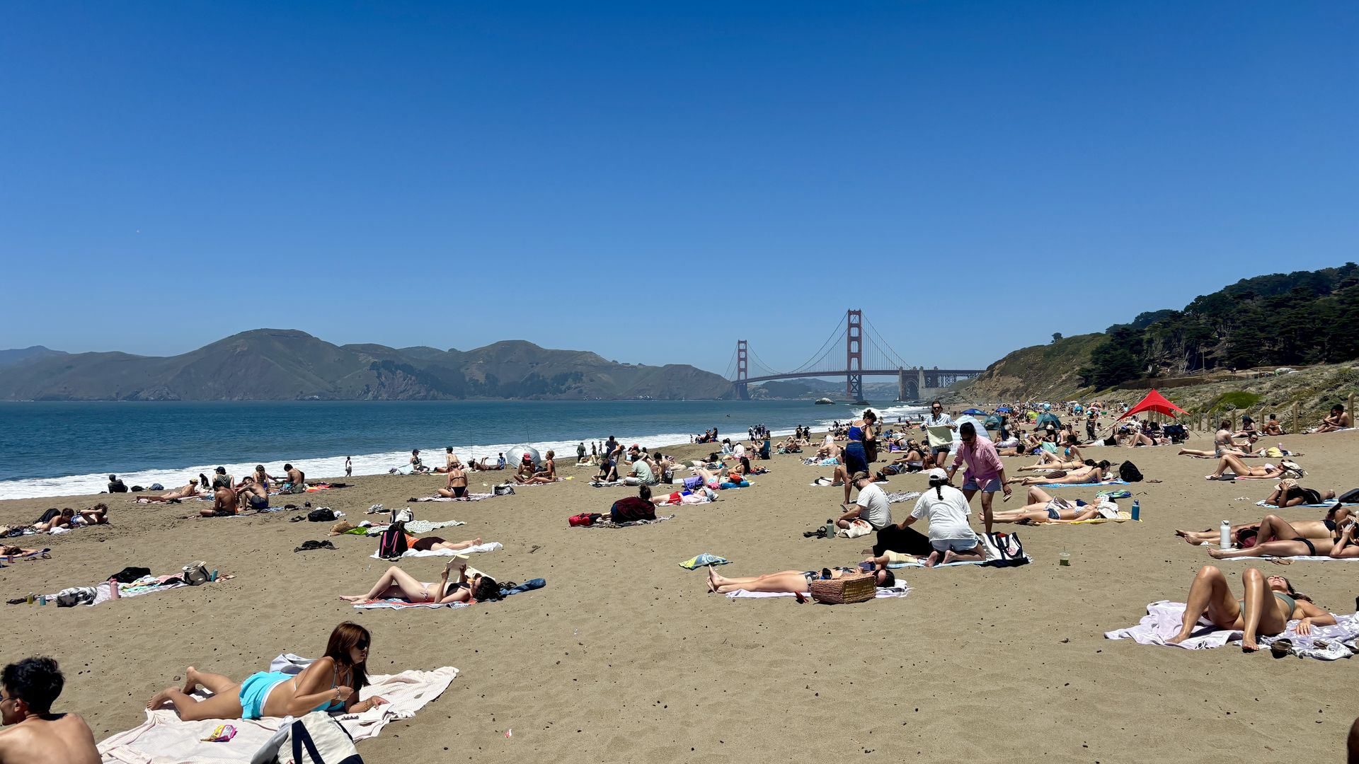 A crowded sandy beach under a bright blue sky, with many sunbathers on towels. In the distance, the red Golden Gate Bridge spans the bay, flanked by hills and trees along the shore.