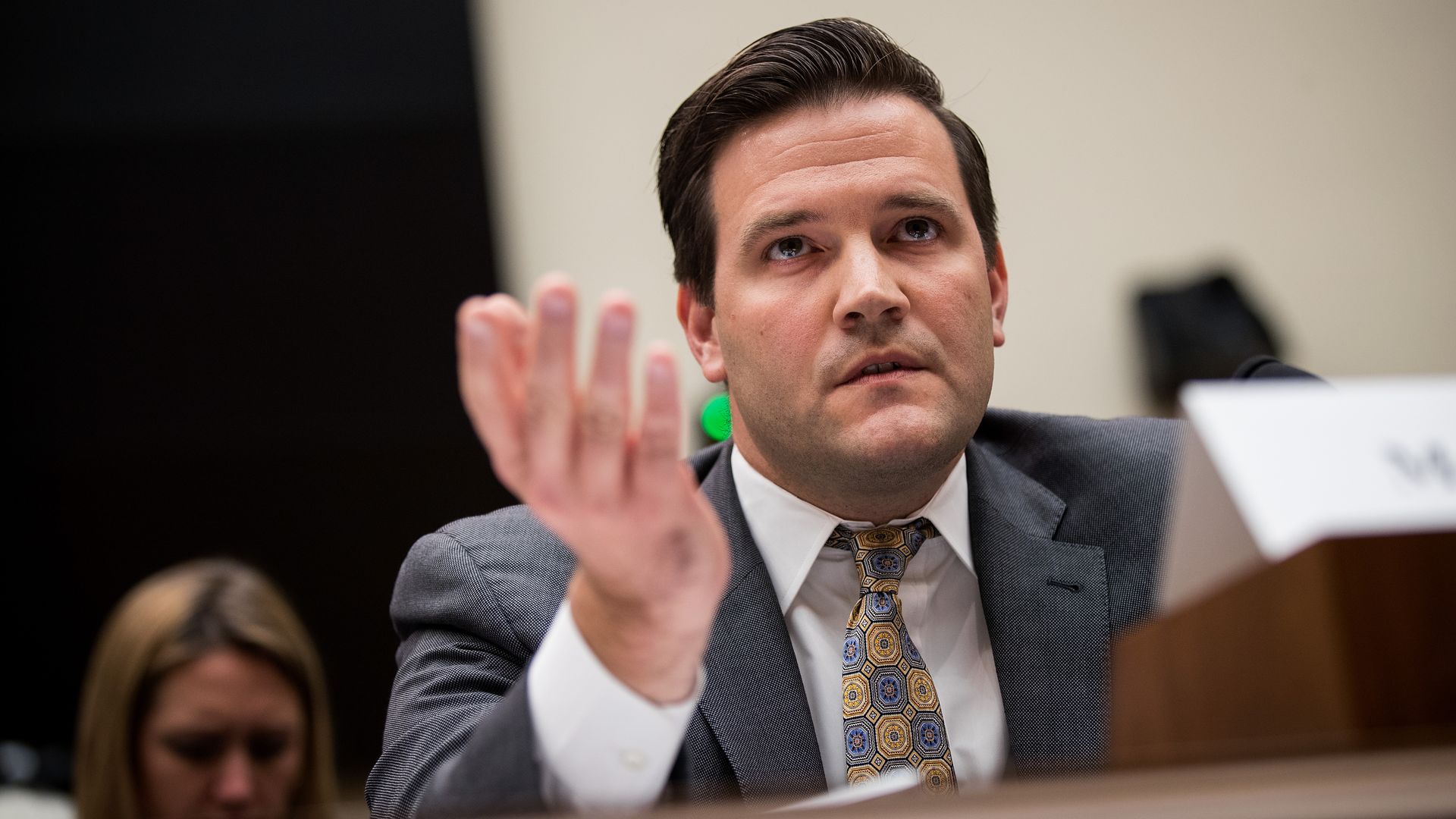 Office of Refugee Resettlement director Scott Lloyd speaking into a microphone at a hearing with one hand in the air