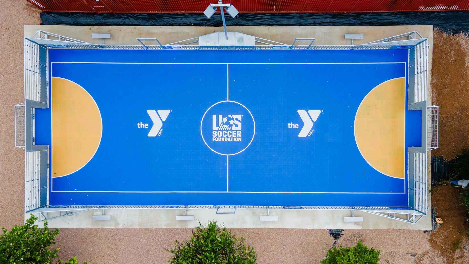 Aerial view of a bright blue outdoor court with white lines. Center circle reads 'SOCCER FOUNDATION' and is flanked by YMCA logos; metal railings frame the court, with beige ground and green trees around.