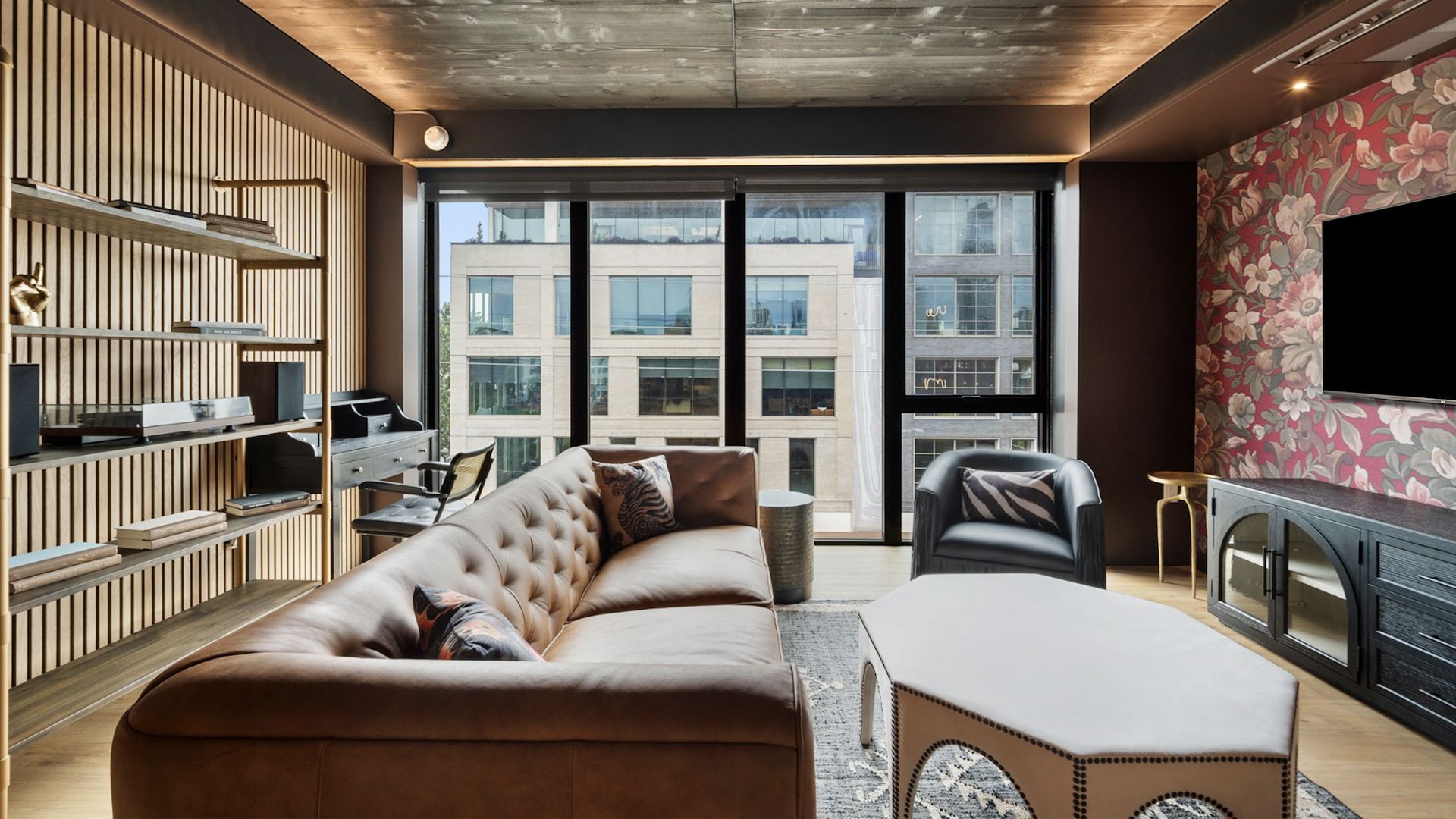 Modern living room with large beige tufted sofa, black armchair with animal print pillows, hexagonal coffee table, floral wallpaper, wood slat wall shelves, and floor-to-ceiling windows.