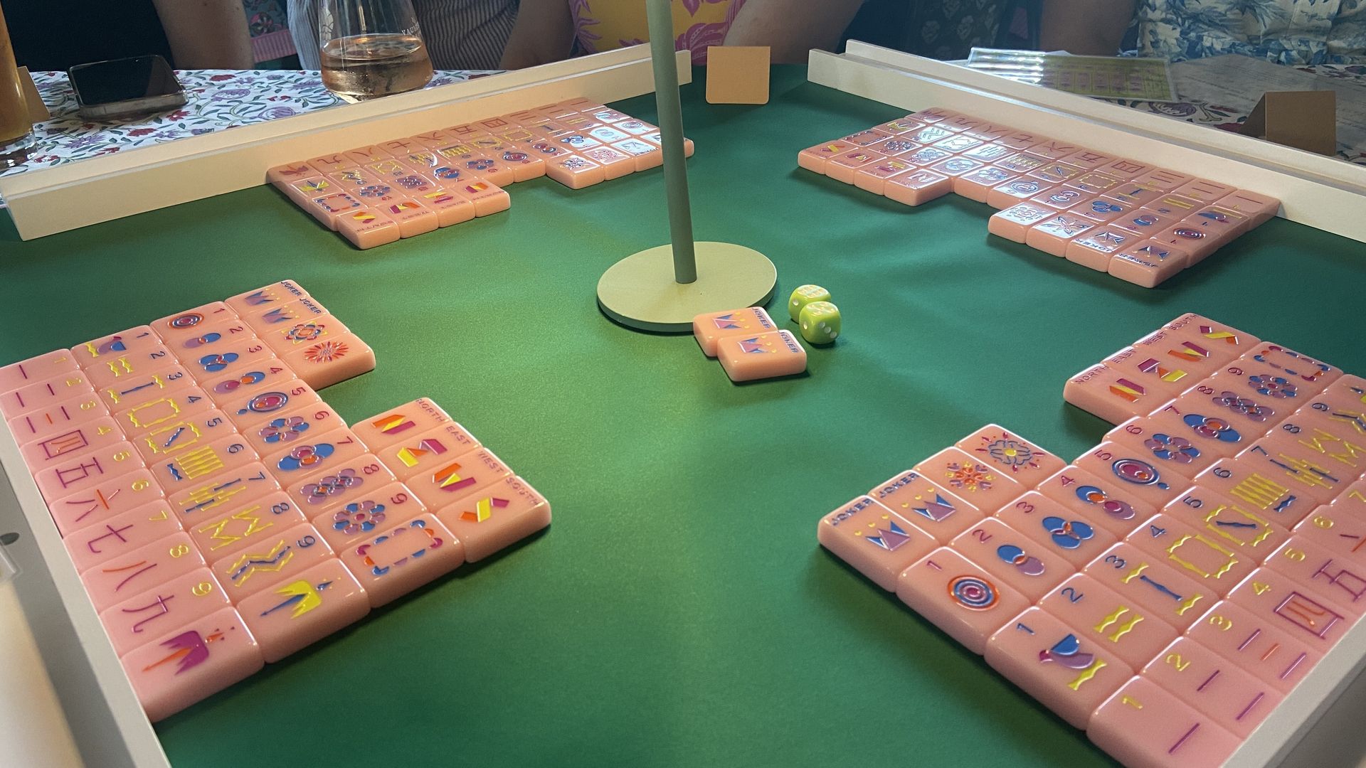 A green playing surface with four sets of pink Mahjong tiles arranged in front of players, a center pole, two green dice, and a table with floral cloth in the background.