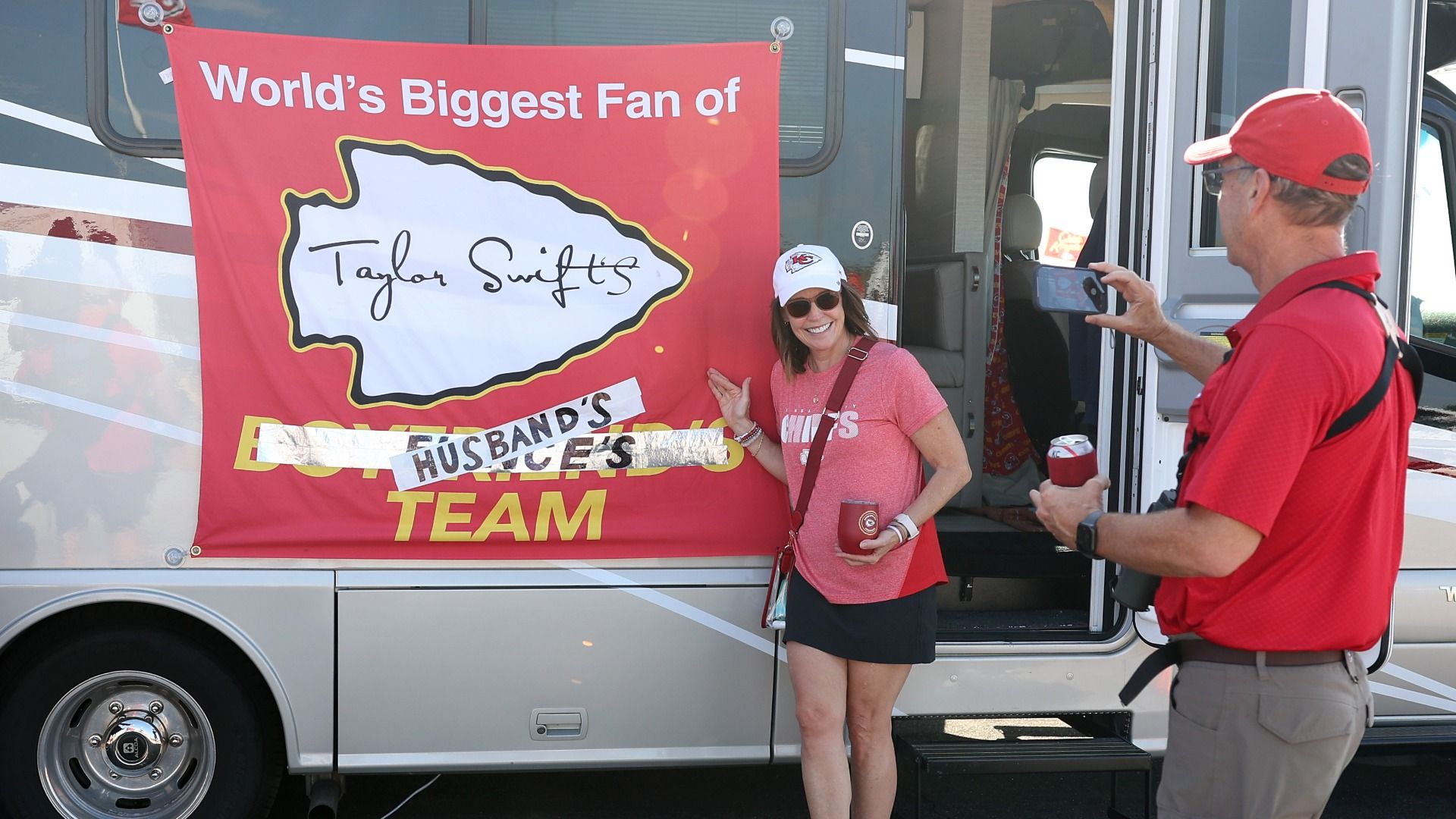 A fan gets her picture taken in front of a bus with a big red sign reading "World's Biggest Fan of Taylor Swift's Husband's Team" with a big Arrowhead logo. 