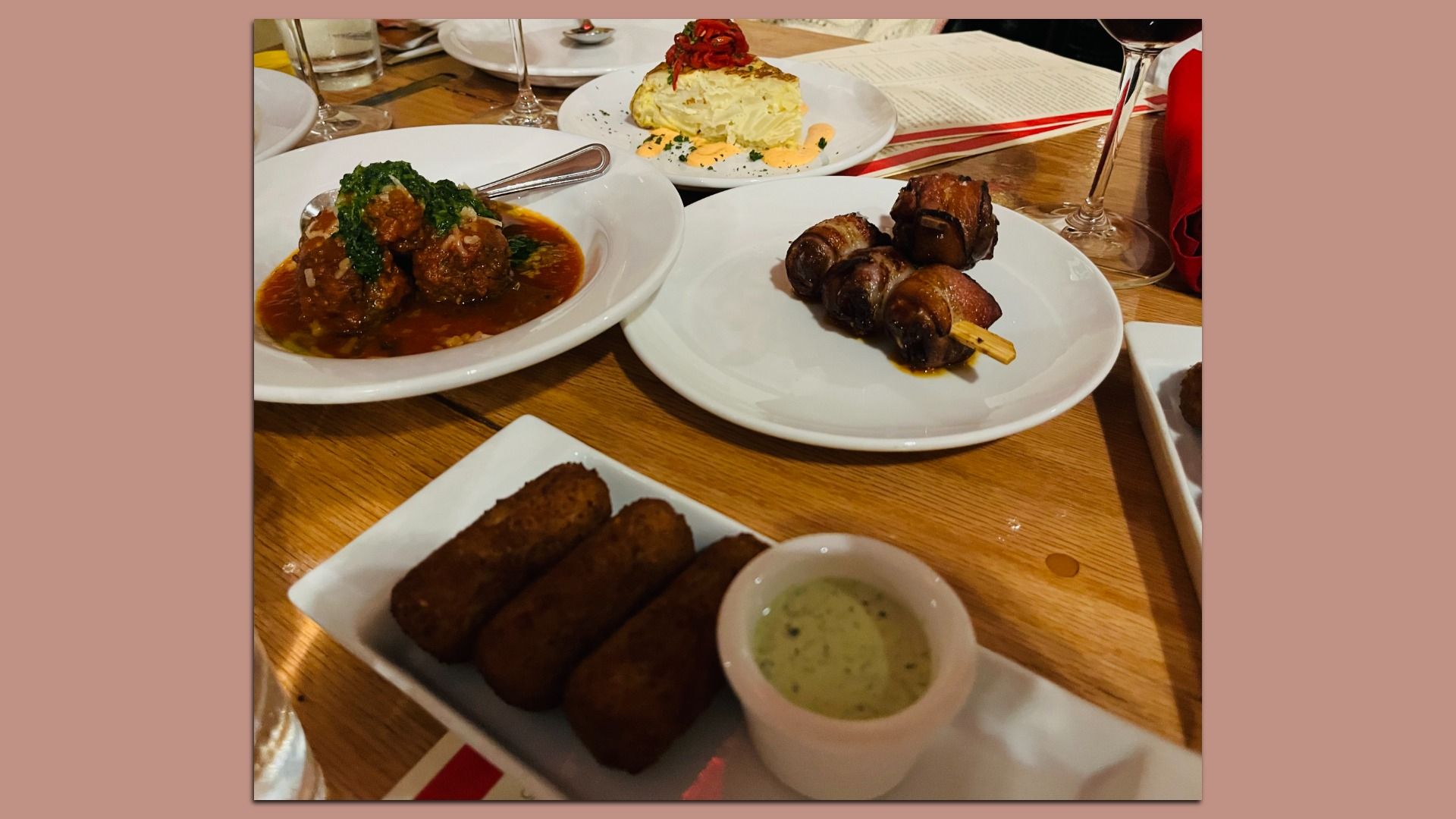 Table with plates of food including meatballs with sauce and herbs, bacon-wrapped mushrooms on a skewer, four fried croquettes with green dipping sauce, and a wedge of potato dish topped with red peppers.