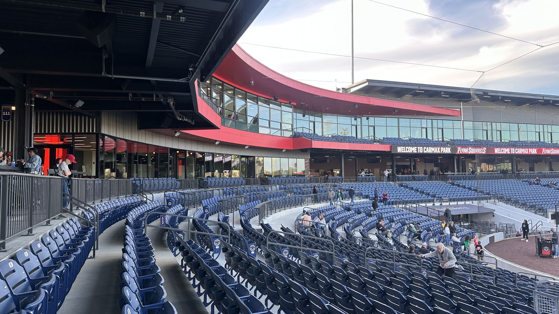 View of a baseball stadium concourse with a curved red exterior above glass-walled levels, rows of blue seats, and a few people walking along the aisles.