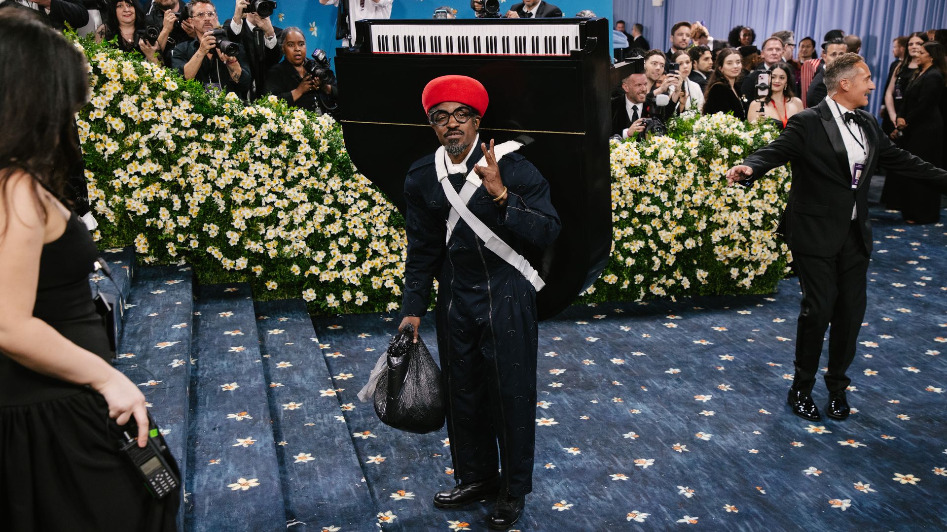 An individual dressed all in black and a red beret hat walks the opening carpet of a gala event with a piano strapped on their back