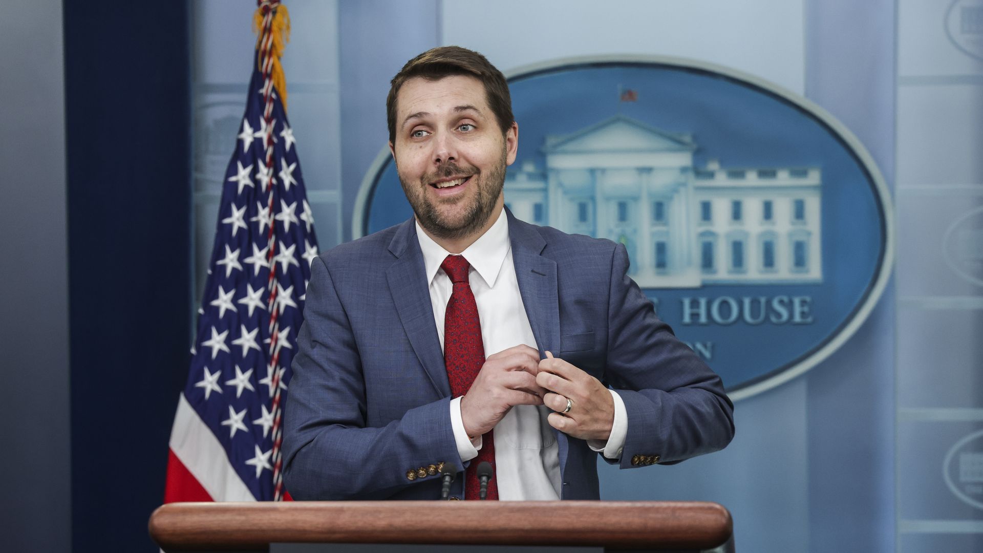 Brian Deese in a red tie and blue suit stands and smiles at the White House lectern