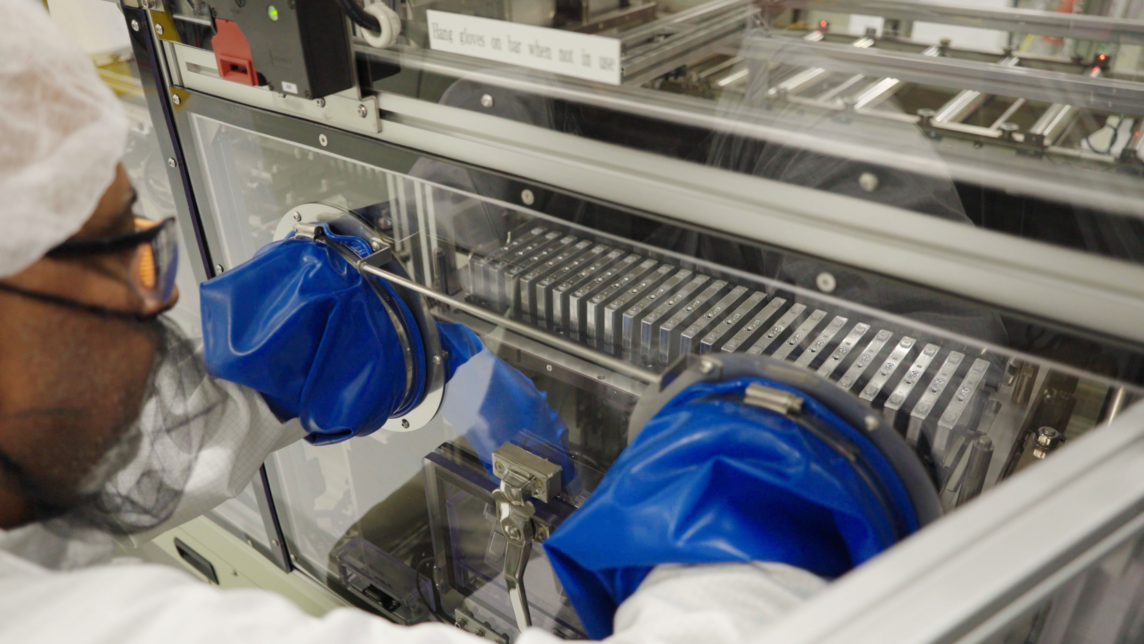 Scientist in protective gear using blue sealed gloves to handle lab equipment inside a transparent containment box in a sterile environment.