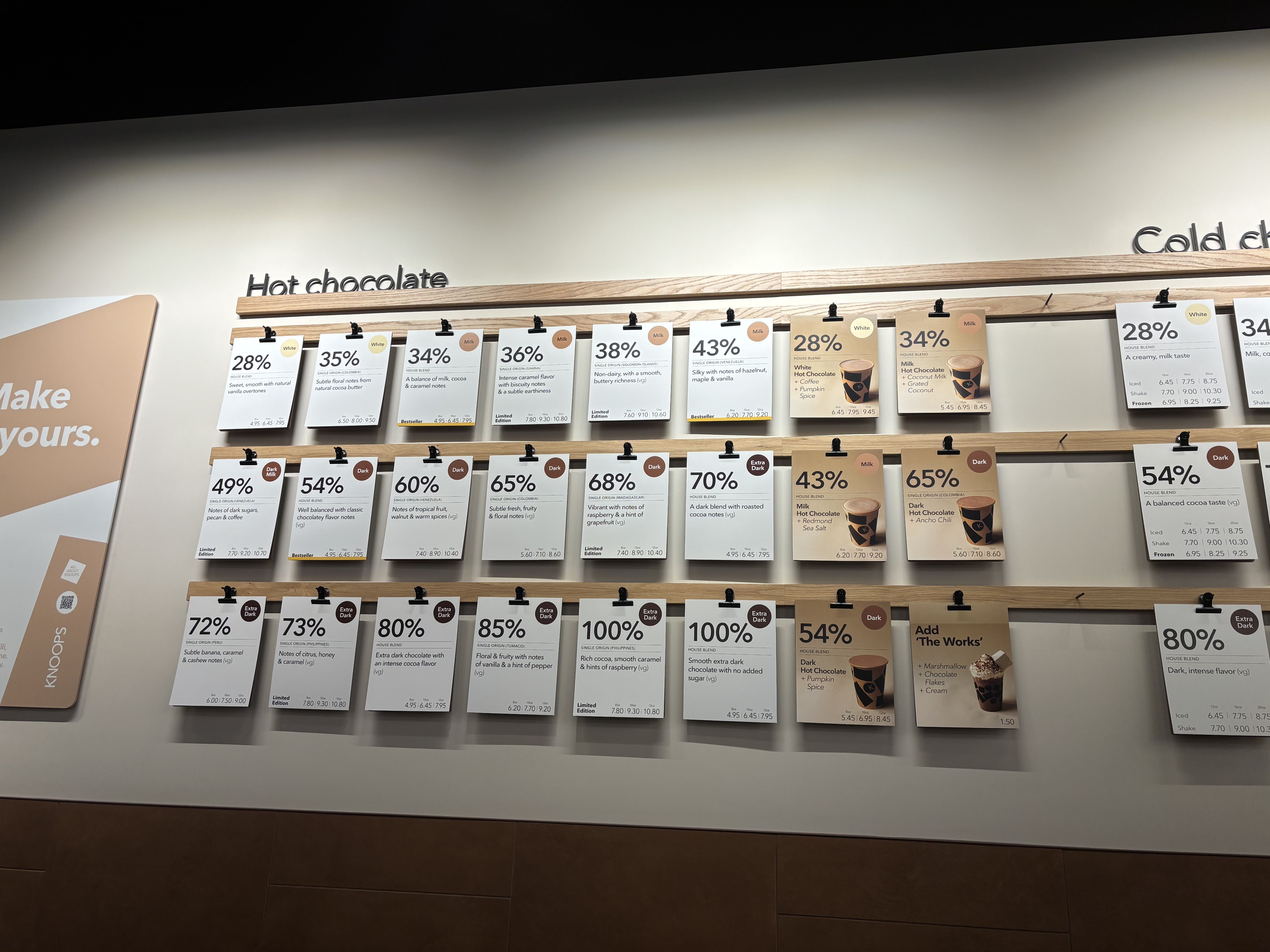 Retail display of chocolate bars on wooden shelves, with a large Hot chocolate sign. Rows of white price cards show percentages (28%, 34%, 36%) and flavor notes against beige walls.