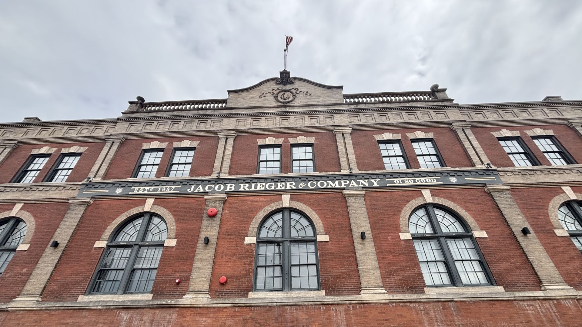 Red brick historic building with tall arched windows, beige stone trim, and a curved central pediment. A small flag waves atop; the façade reads "JACOB RIEGER & COMPANY".