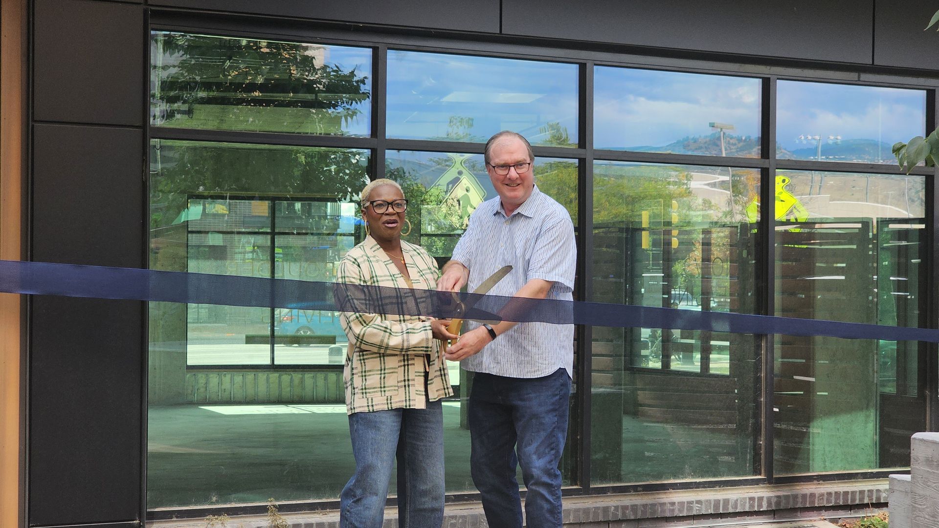 Mawa McQueen and Boulder Mayor Aaron Brockett stand in front of a glass building holding a large pair of scissors cutting a dark blue ribbon for a ribbon-cutting ceremony.