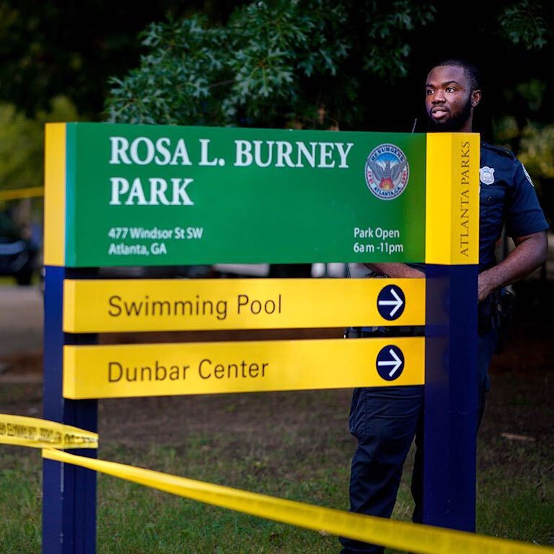 police officers standing next to park entry sign with crime tape wrapped around it
