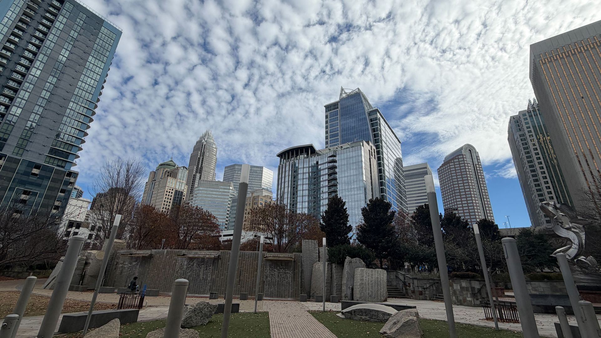 City park with green grass and stone sculptures in foreground, surrounded by tall modern glass and concrete skyscrapers under a cloudy blue sky.