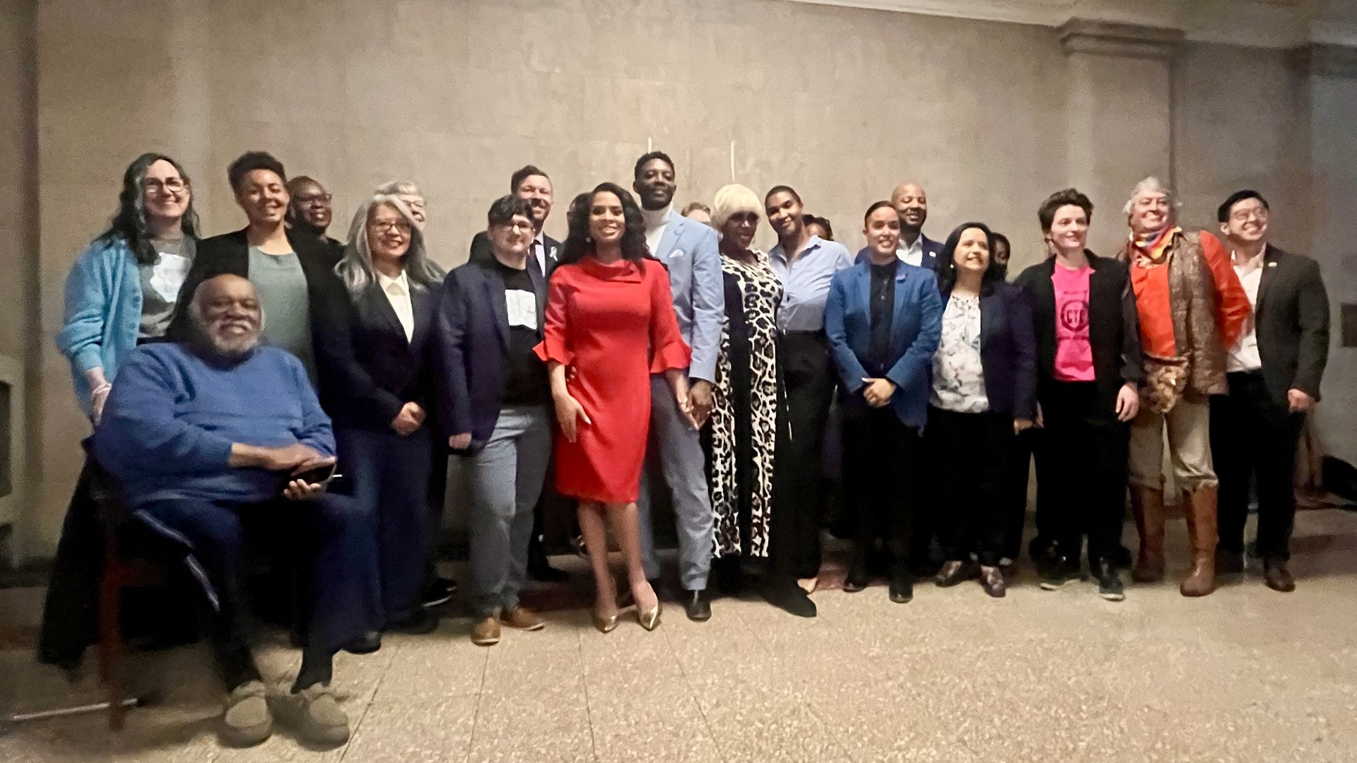 People standing in a line in front of a brick wall with one man sitting on a chair on the end.