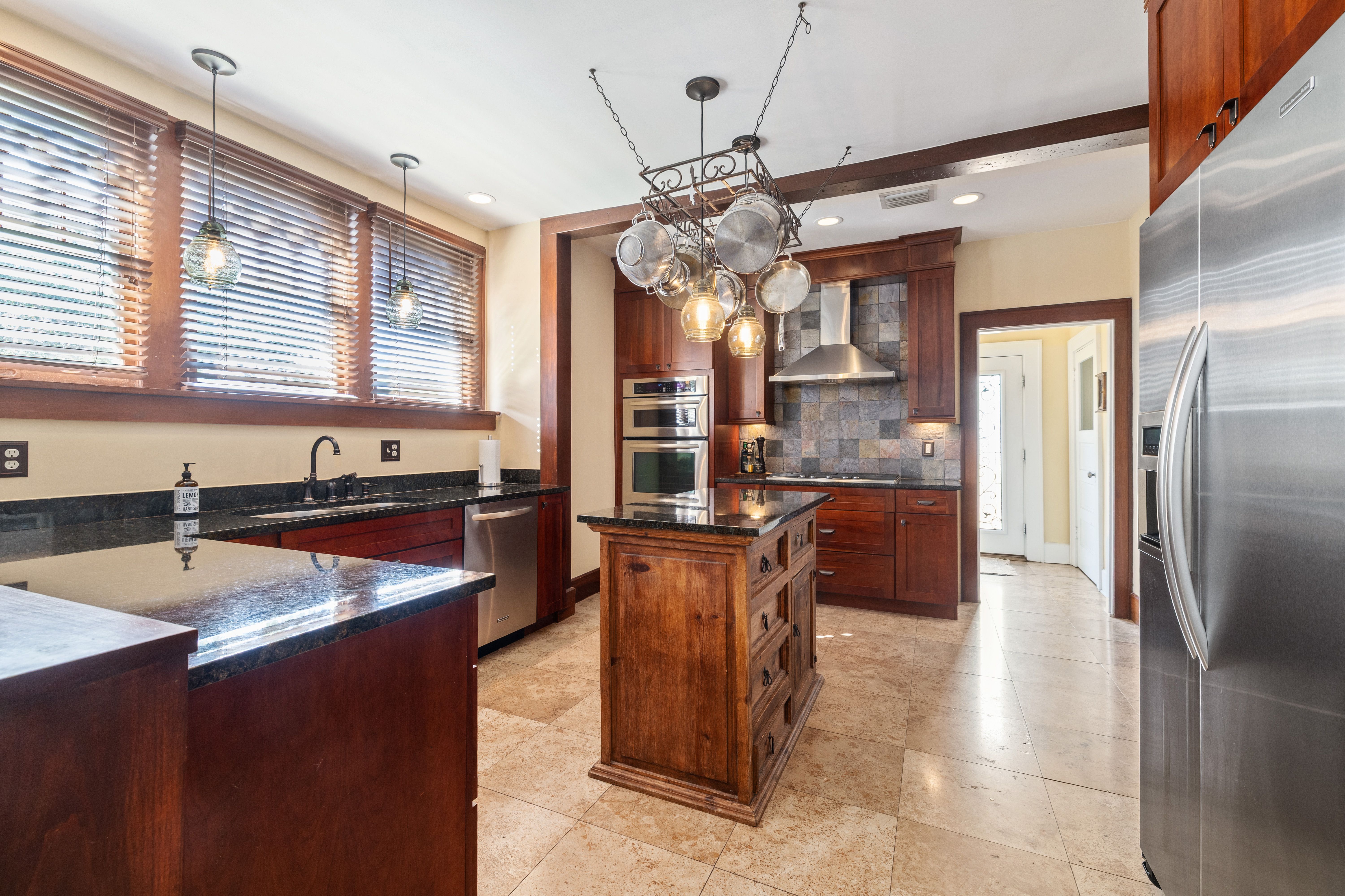 Bright kitchen with dark granite countertops, wooden cabinets, stainless steel appliances, hanging pots, and beige tiled floor. Large windows with wooden blinds let in sunlight.