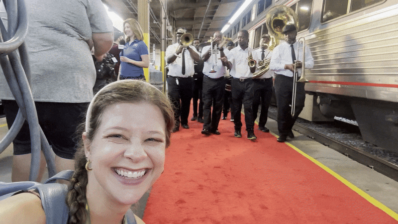A smiling woman takes a selfie on a red carpet at a train station while a brass band in white shirts and black pants plays behind her next to a silver train.