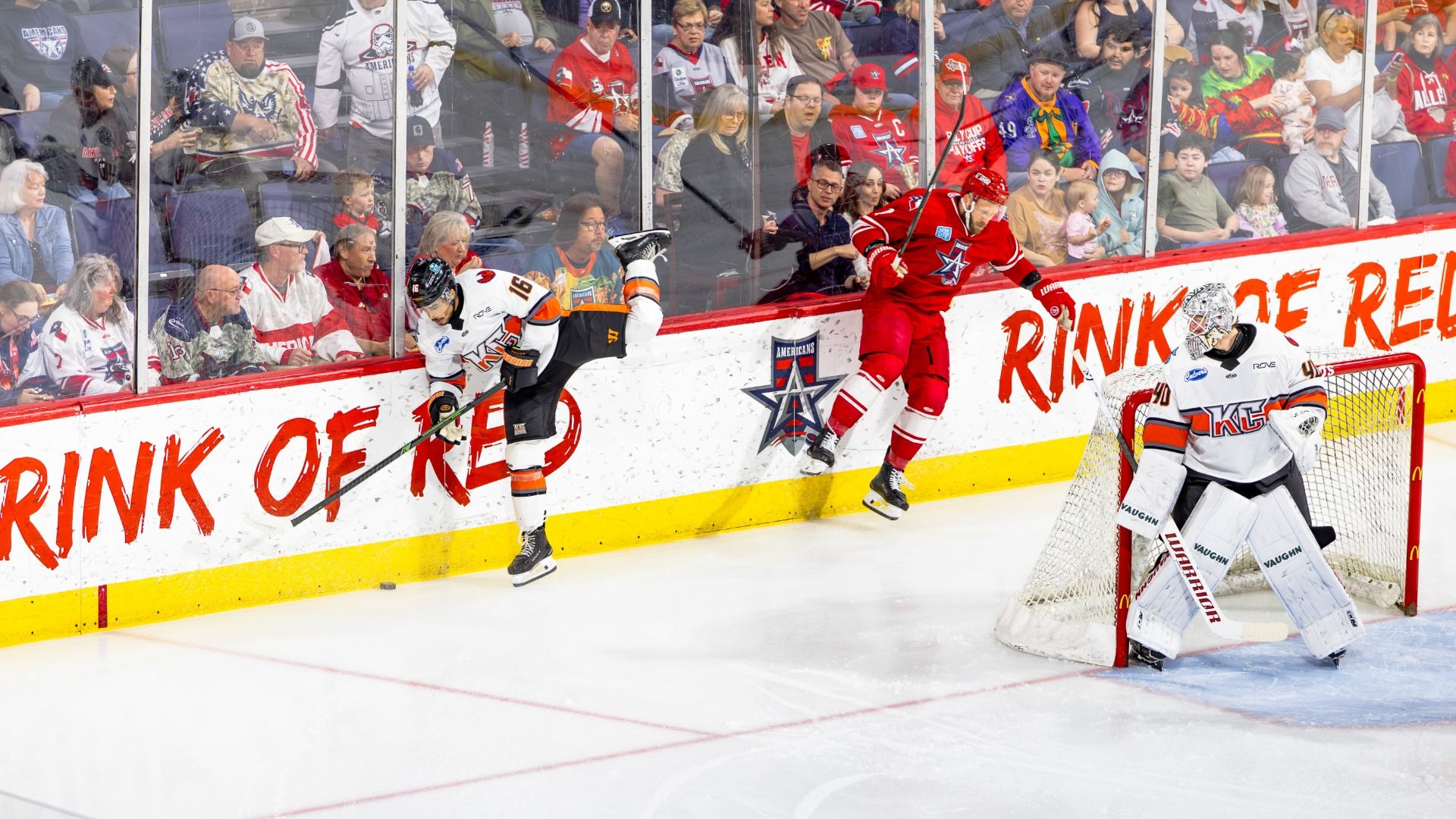 Red-clad hockey player skates along the boards past a white-uniform opponent while the goalie in white guards the net; a packed crowd watches through glass panels.