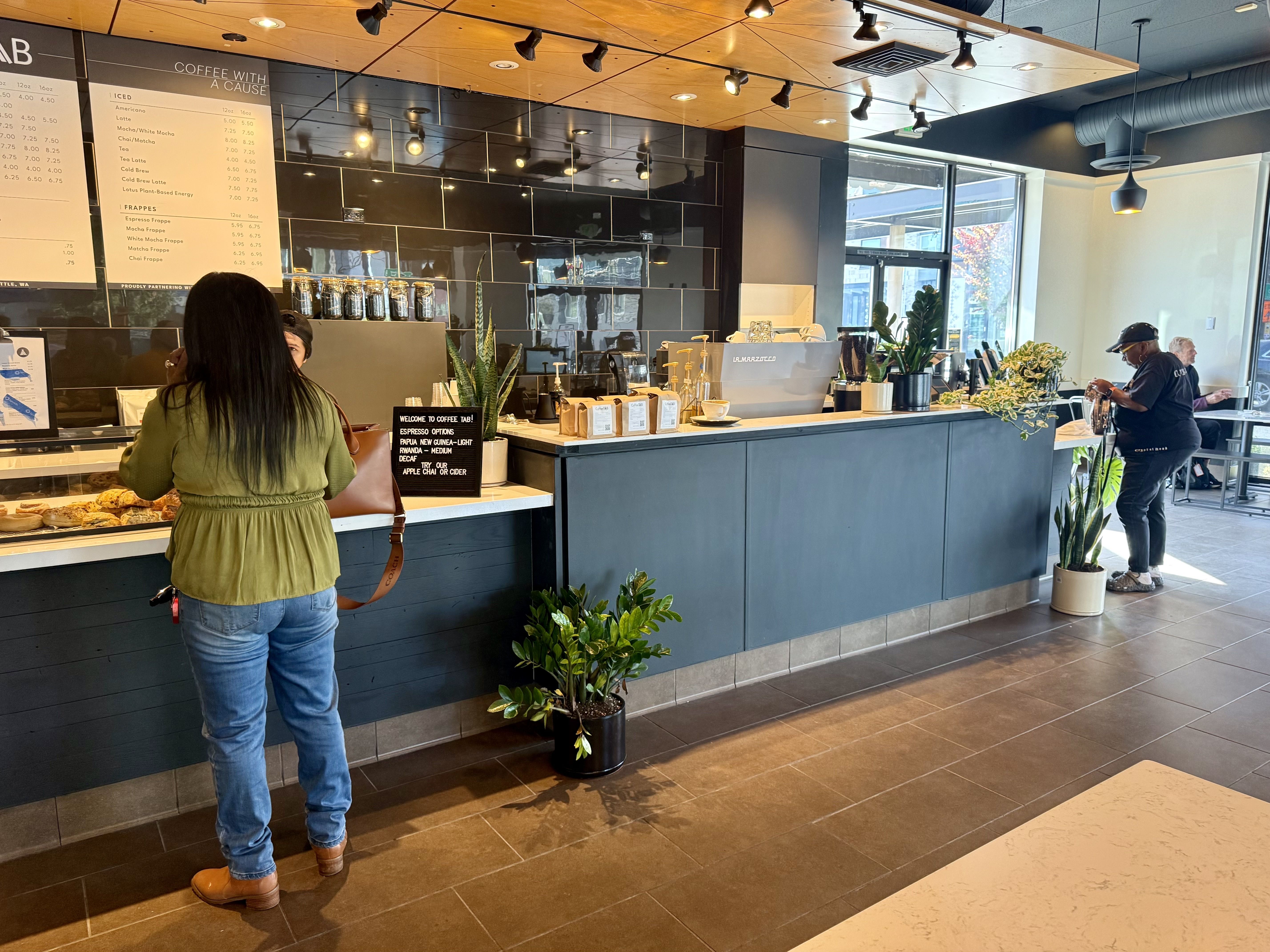 Inside a modern cafe with dark tile walls and a long counter. A pastry display sits near the front; a customer in a green jacket stands left, a worker with a cap at the back, by large windows and plants.
