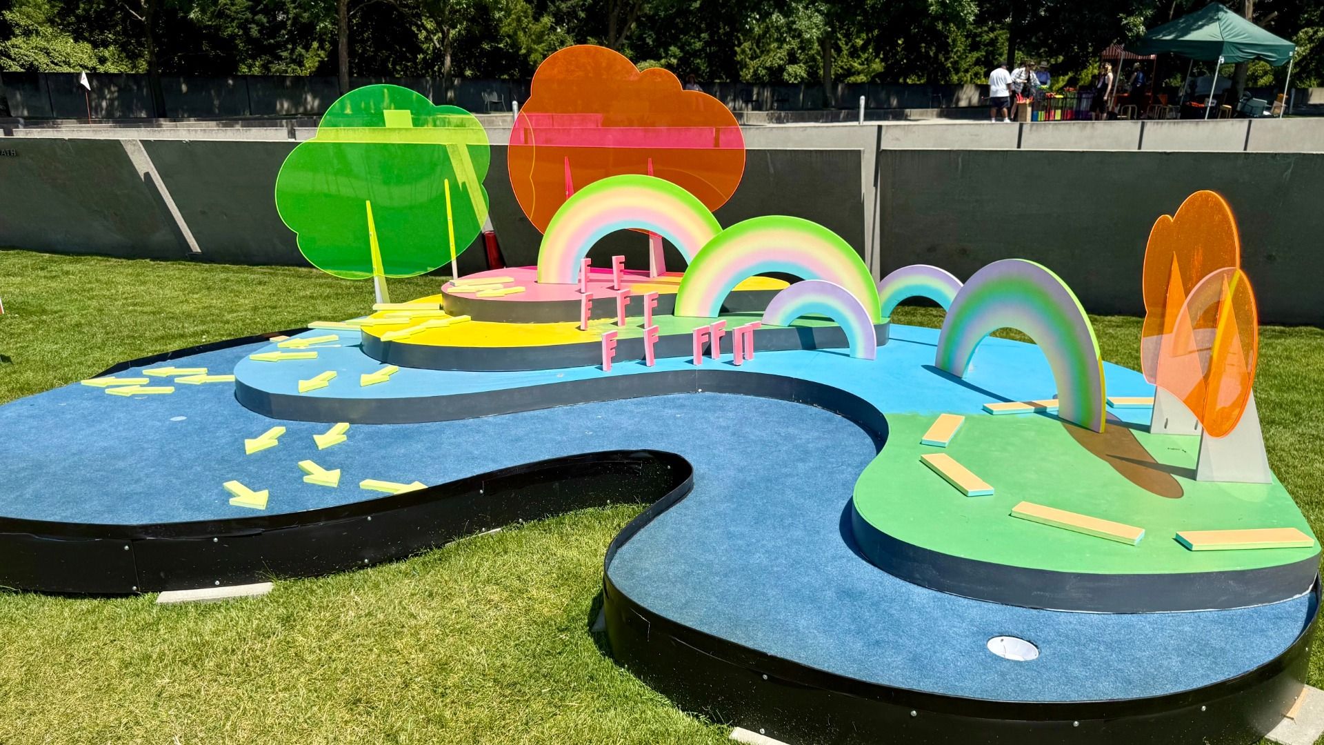 Vibrant outdoor sculpture with oversized neon green and orange cloud shapes, pastel rainbow arches on blue platforms, yellow stepping blocks, and a grassy field; people near a green tent in the background.