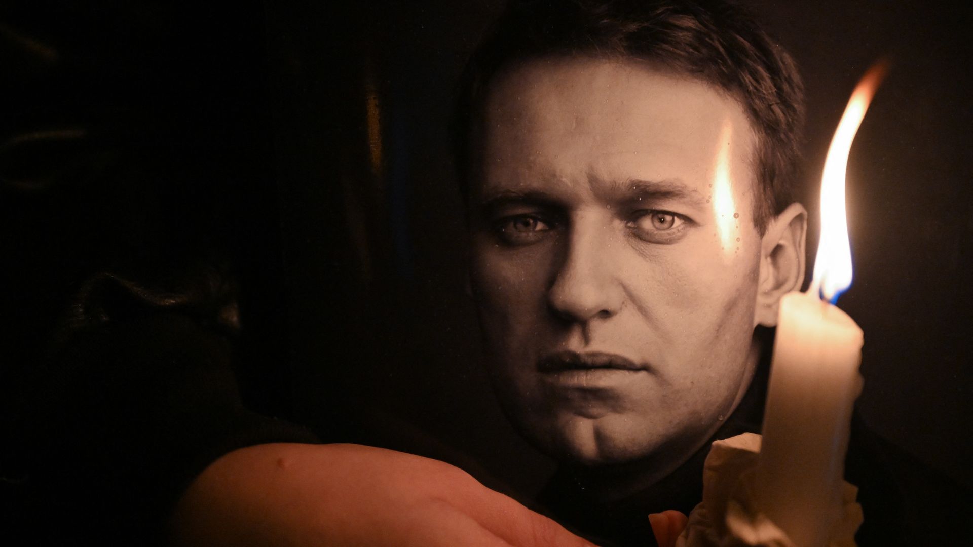 Person holding a lit white candle with a cloth in dim lighting, next to a close-up black-and-white photo of the late Kremlin critic Alexy Navalny's serious face.