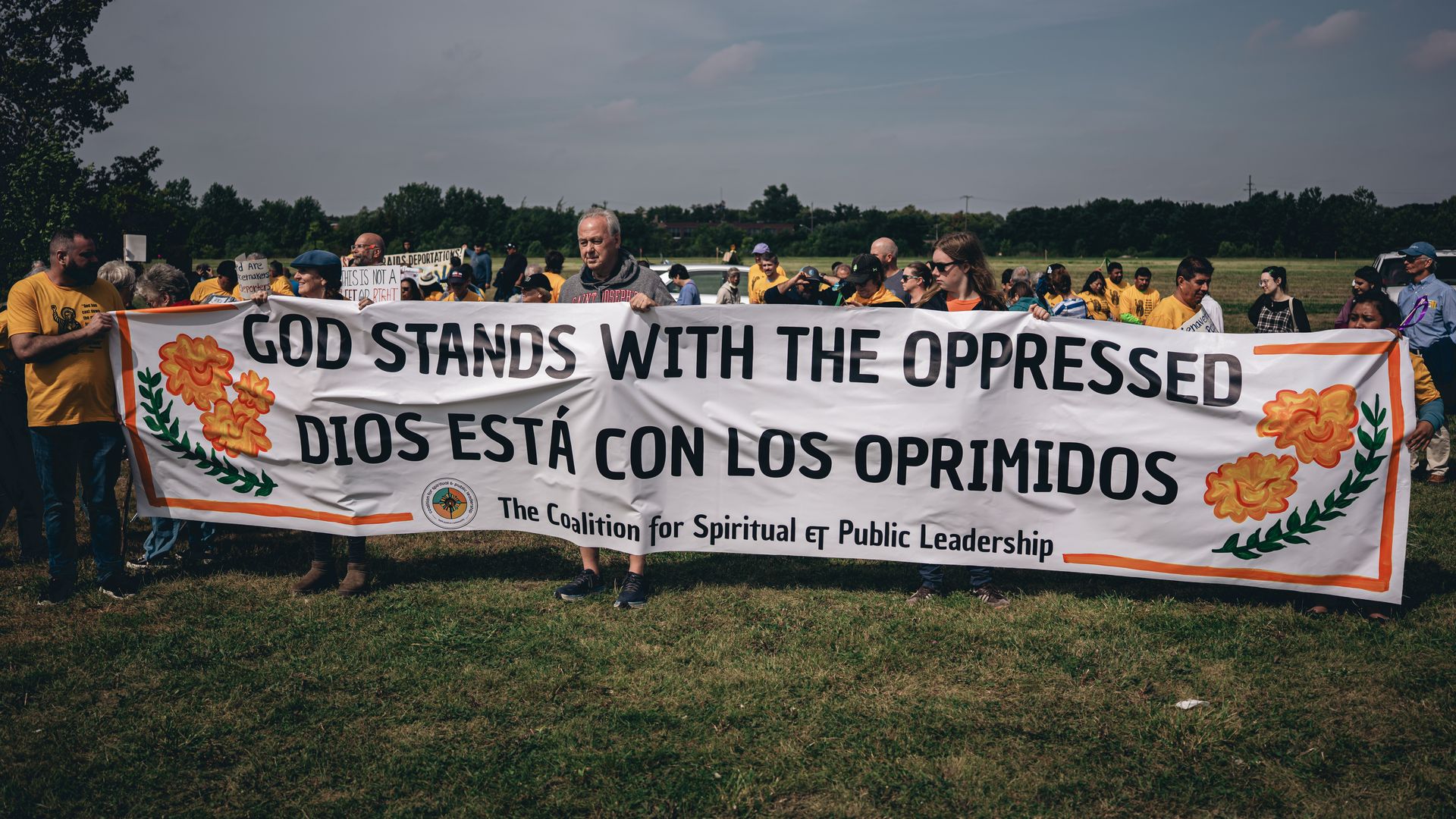 A group of people holding a large banner outdoors that reads "God stands with the oppressed / Dios está con los oprimidos" with orange flowers decorating the edges.