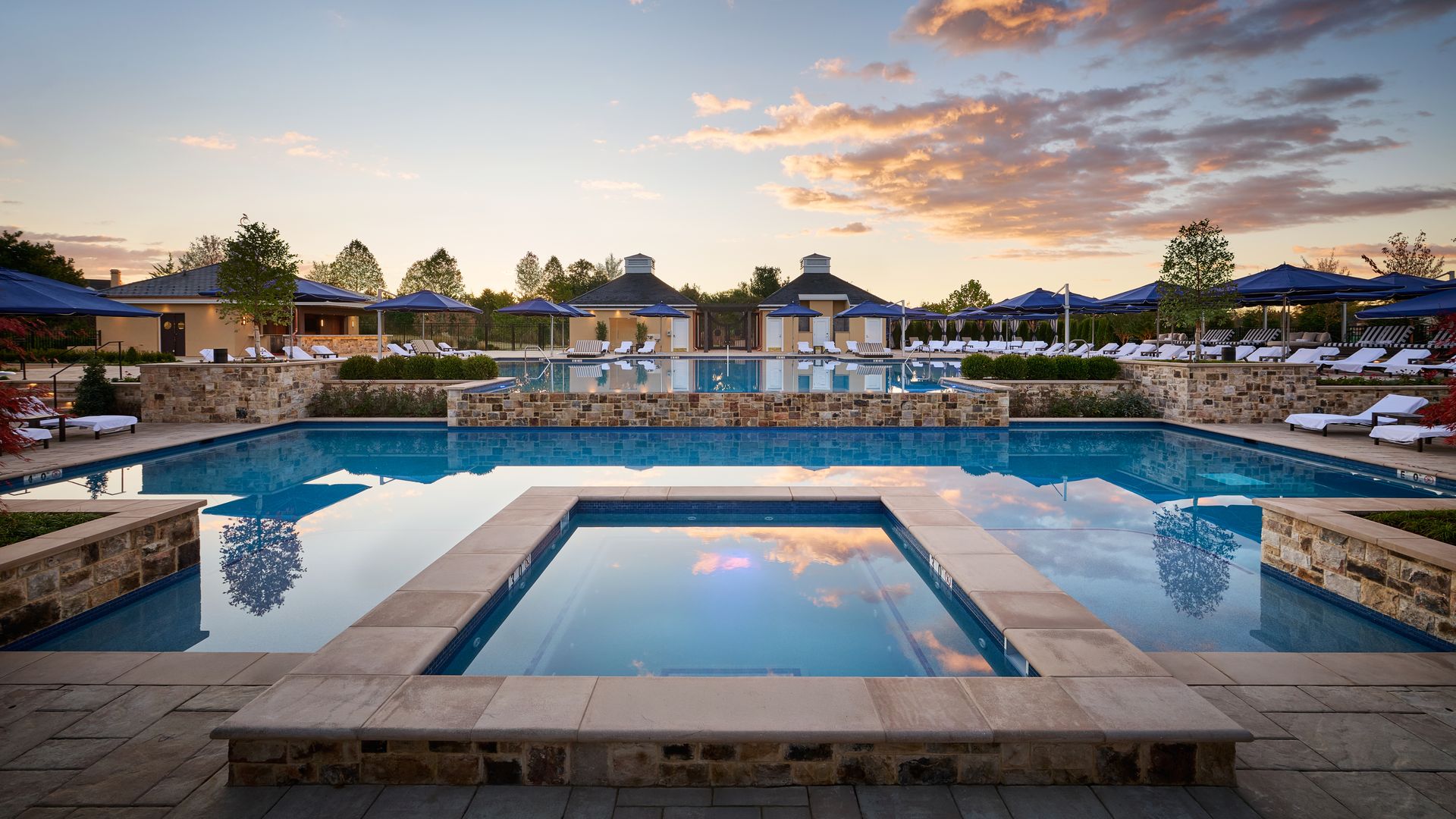 Sunset over a resort pool complex with blue-tiled pools, stone decks and walls, white lounge chairs under blue umbrellas, and calm water reflecting the orange sky and surrounding trees.