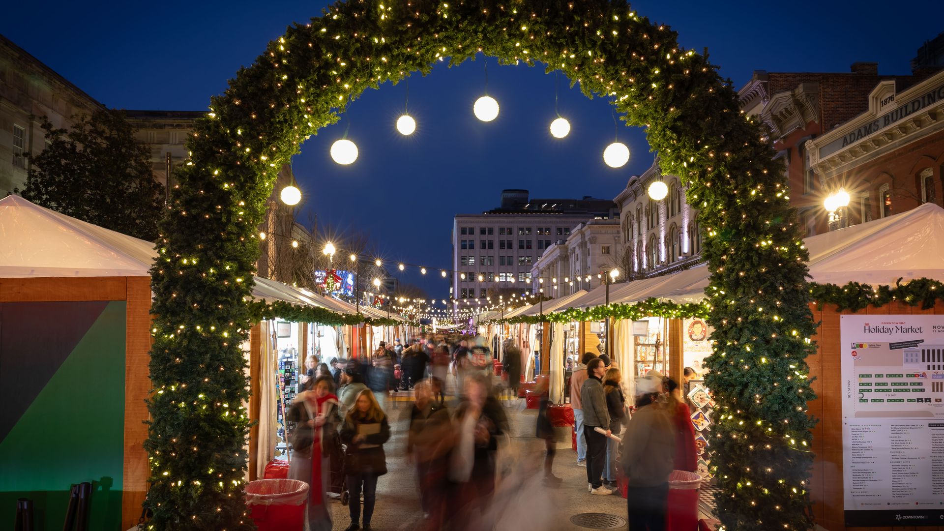 Crowded evening holiday market with illuminated greenery arch, hanging globe lights, festive booths, and people walking under clear dark blue sky in a city street.