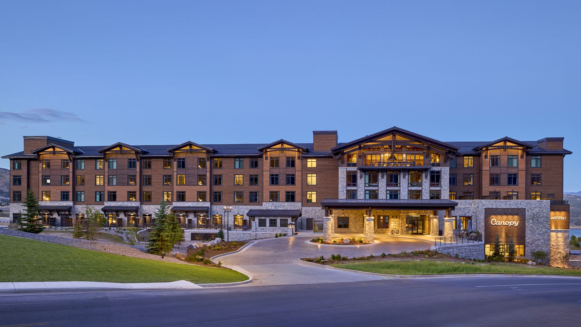 Wide view of a large brown brick and stone hotel with timber-framed, peaked roofs; warmly lit windows, a stone entrance canopy, circular driveway, landscaped grounds, and a blue evening sky.