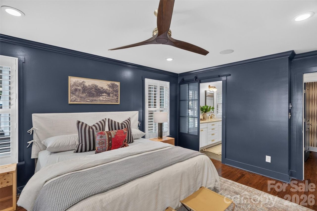 Bedroom with navy walls, white upholstered bed, striped and red accent pillows. Dark wood floor, ceiling fan, framed art, and a glass-front doorway to a bright bathroom with white cabinets.