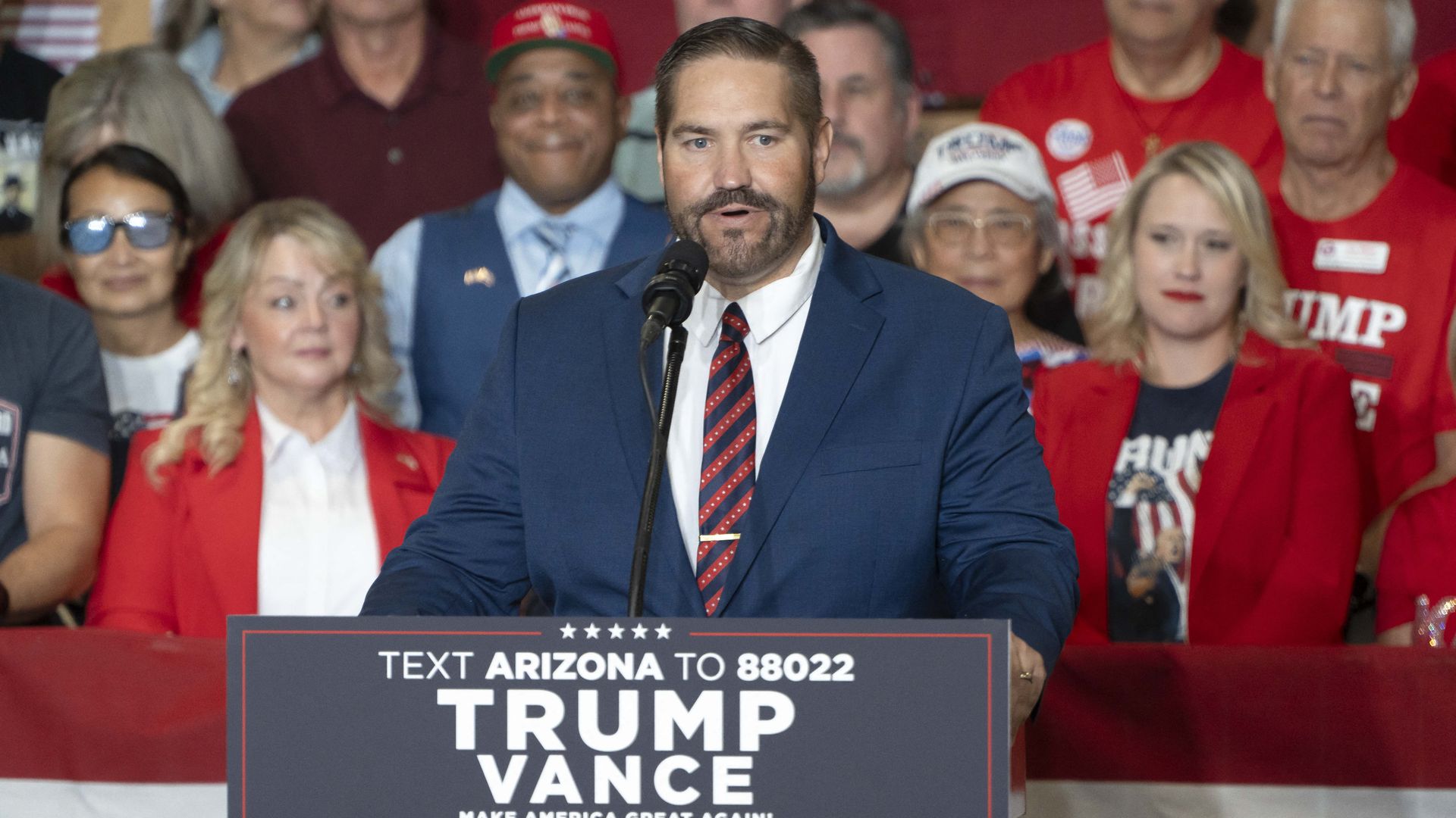 A man in a blue suit speaks at a podium with a sign reading "Text Arizona to 88022 TRUMP VANCE Make America Great Again! 2024". People stand behind him, some wearing red shirts.