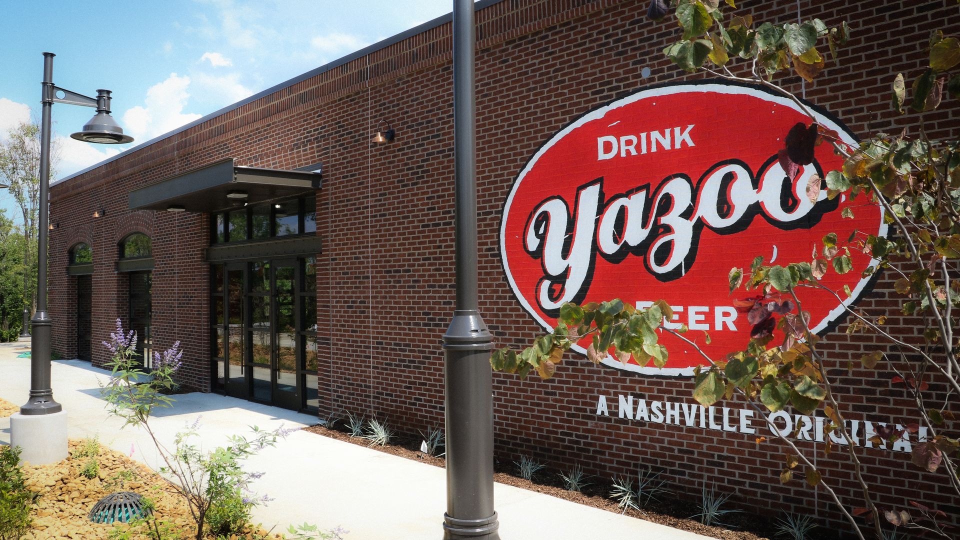 Brick building with a large red circular Coca-Cola sign on the wall reading "Drink Coca-Cola." Glass entrance, black awning, streetlamps, and landscaped foreground under a blue sky.
