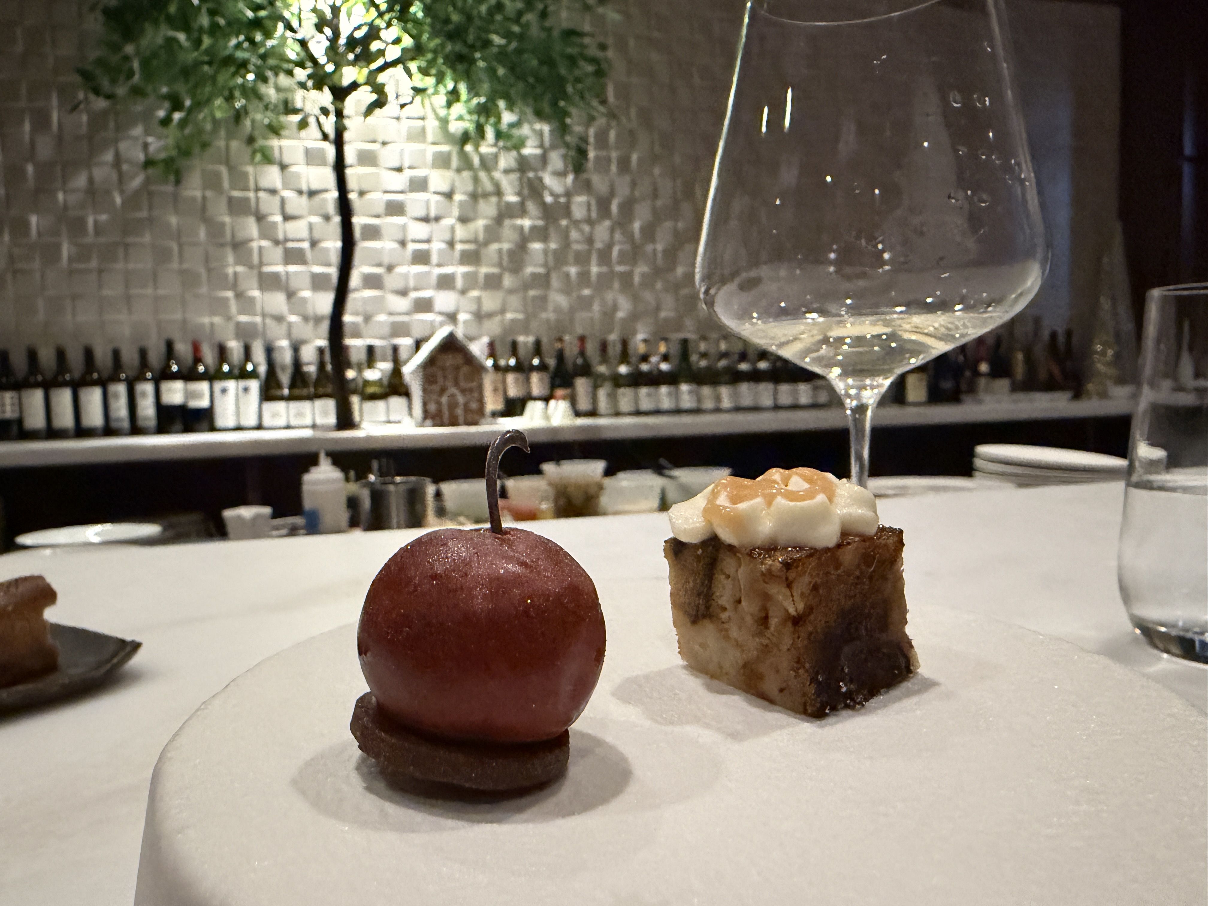 Two small gourmet desserts on a white plate at a bar with wine bottles in the background; one looks like a shiny red sphere with a stem, the other a small square topped with cream.