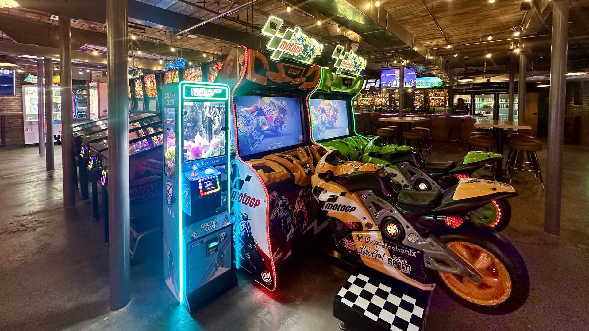 Interior of an arcade bar with colorful MotoGP motorcycle racing arcade games, a claw machine, string lights on a wooden ceiling, and a bar area with stools and TVs.