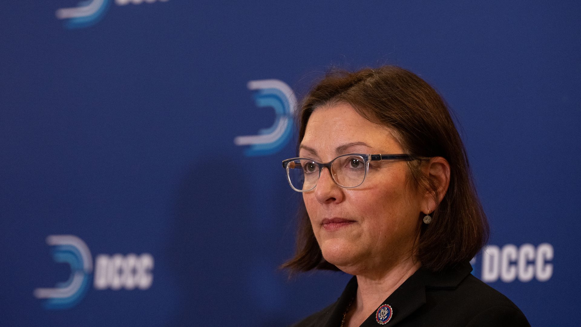 Woman with brown hair and glasses wearing a dark blazer and circular lapel pin, standing in front of a blue backdrop with white DCCC logos.