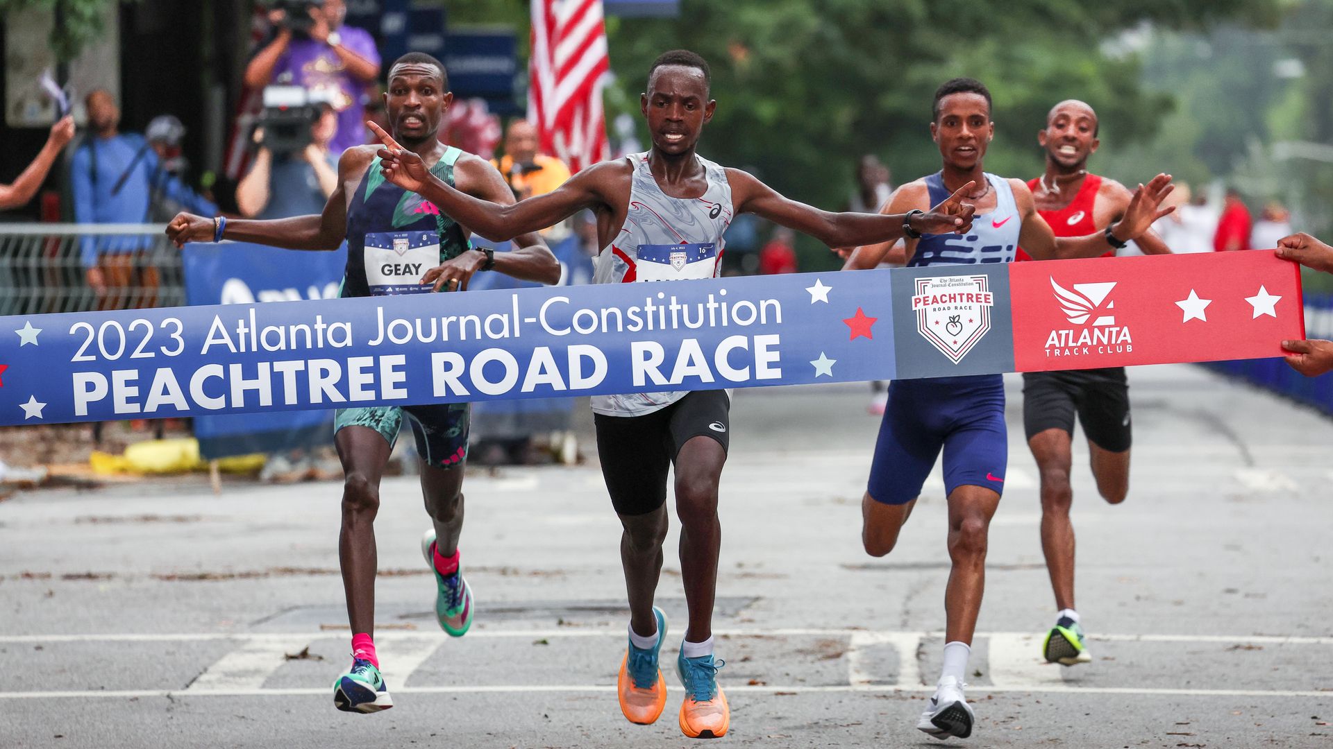 A runner stretches his arms wide as he runs through a finish-line ribbon that says “Peachtree Road Race”