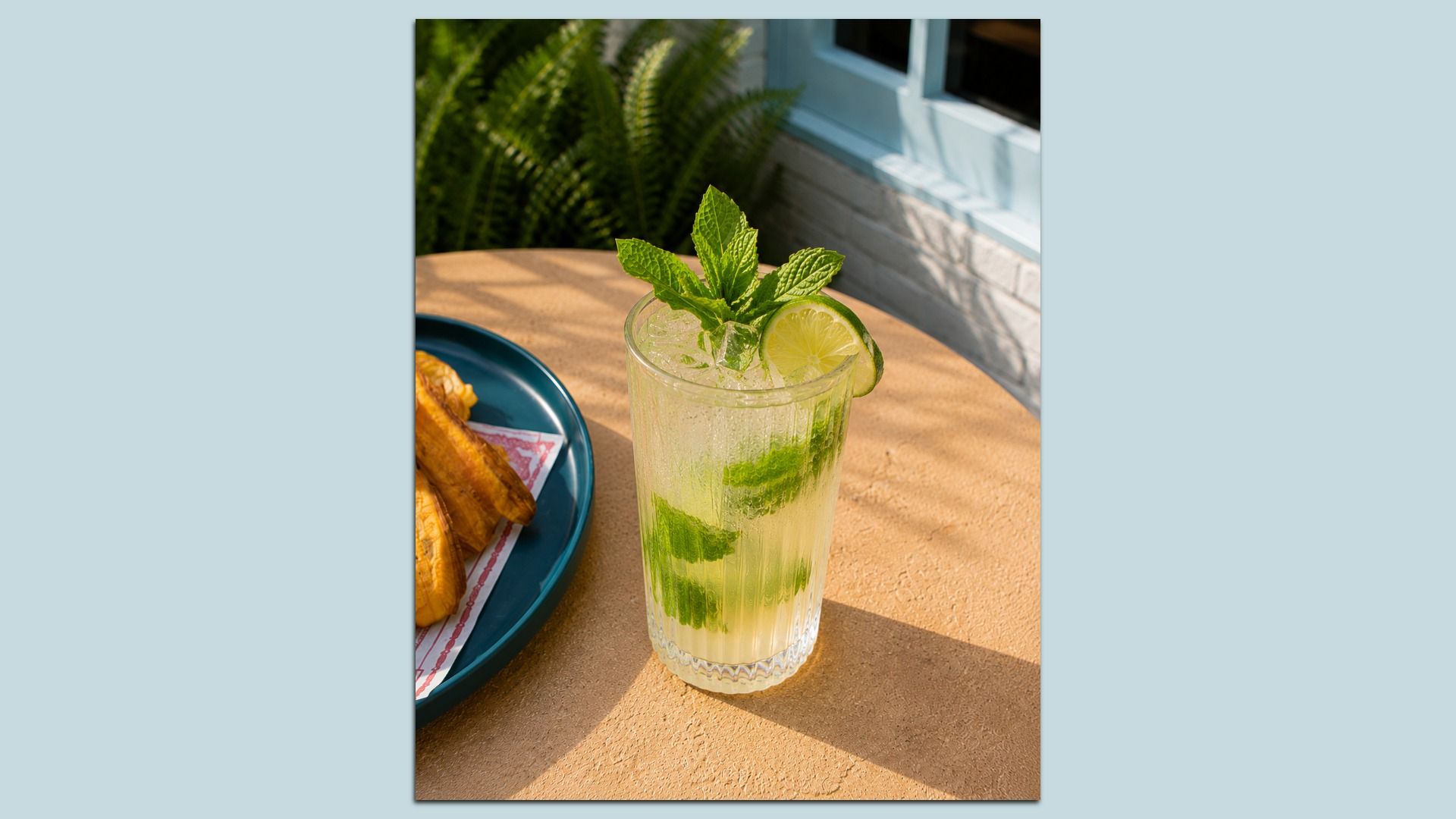 Glass of mojito with mint and lime wheel on a sunlit outdoor table; a blue plate with pastries sits nearby, with green plants and a light blue building in the background.