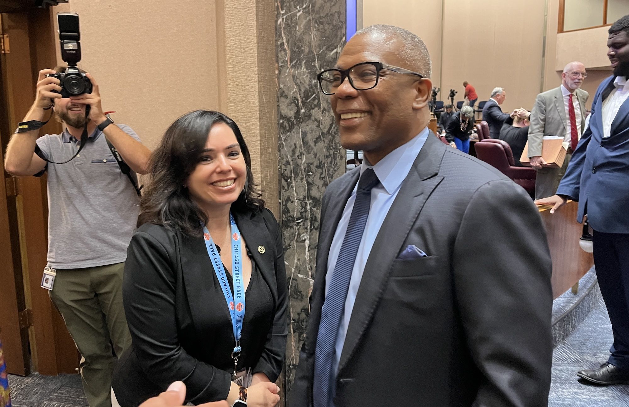 Cristina Pacione-Zayas smiling next to Walter Burnett while a photographer captures the moment. Others in suits in background.
