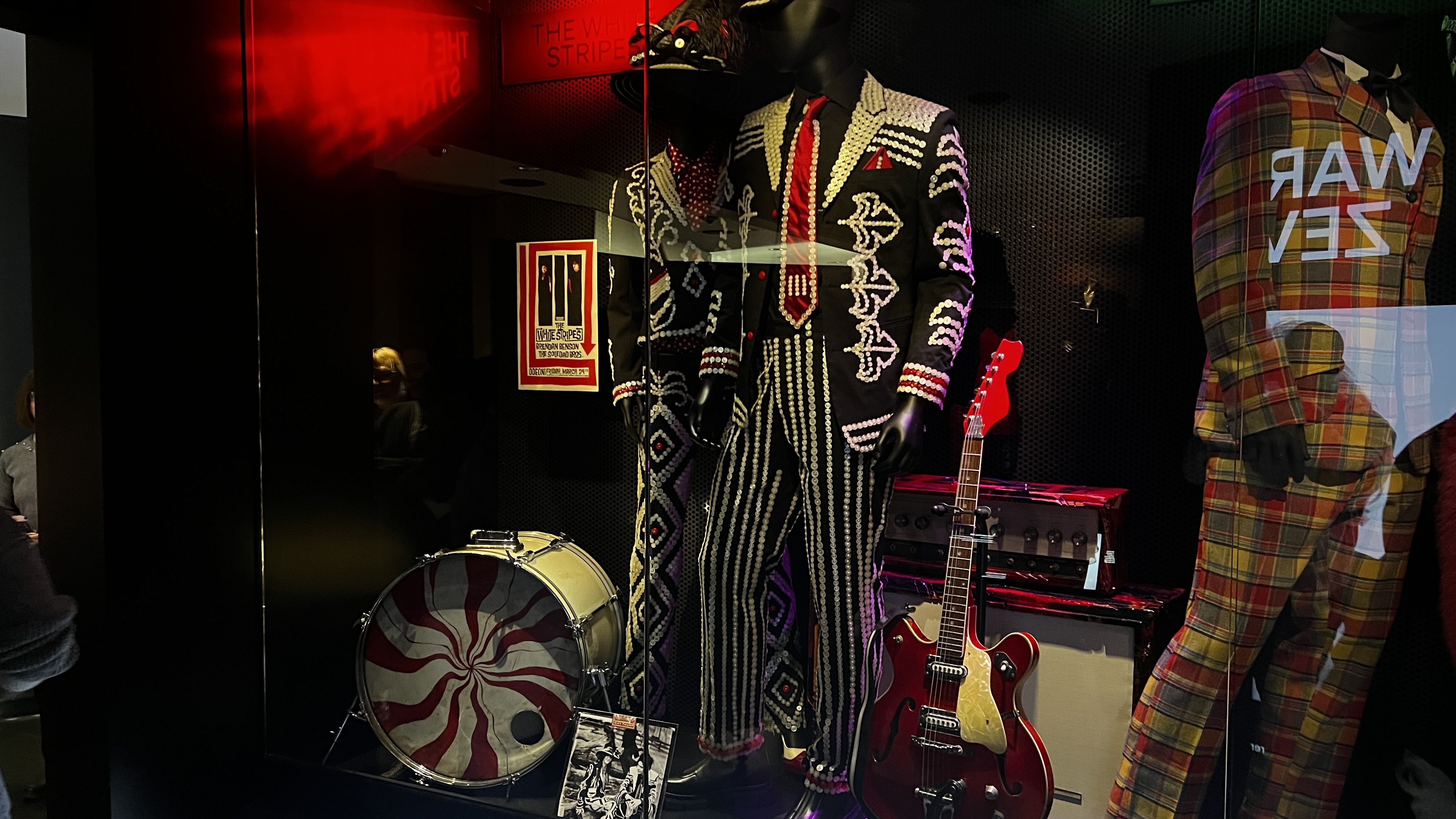 Display of vintage band outfits with black mannequins dressed in black and white patterned suits, a red and yellow plaid suit, a red electric guitar, and a drum with red spiral design.