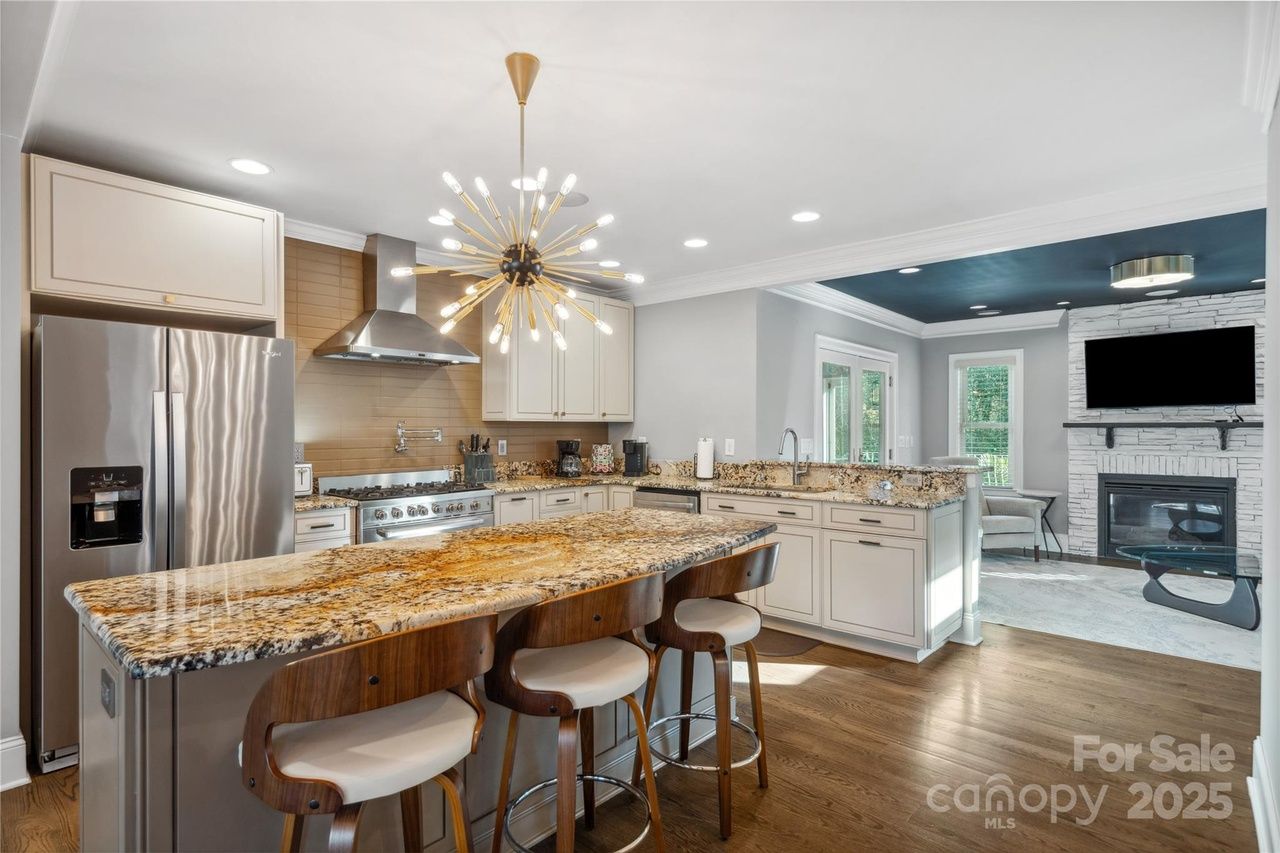 Modern kitchen with granite island, three wood and white stools, stainless steel refrigerator and stove, beige cabinets, stylish chandelier, hardwood floors, adjacent living room with stone fireplace and TV.