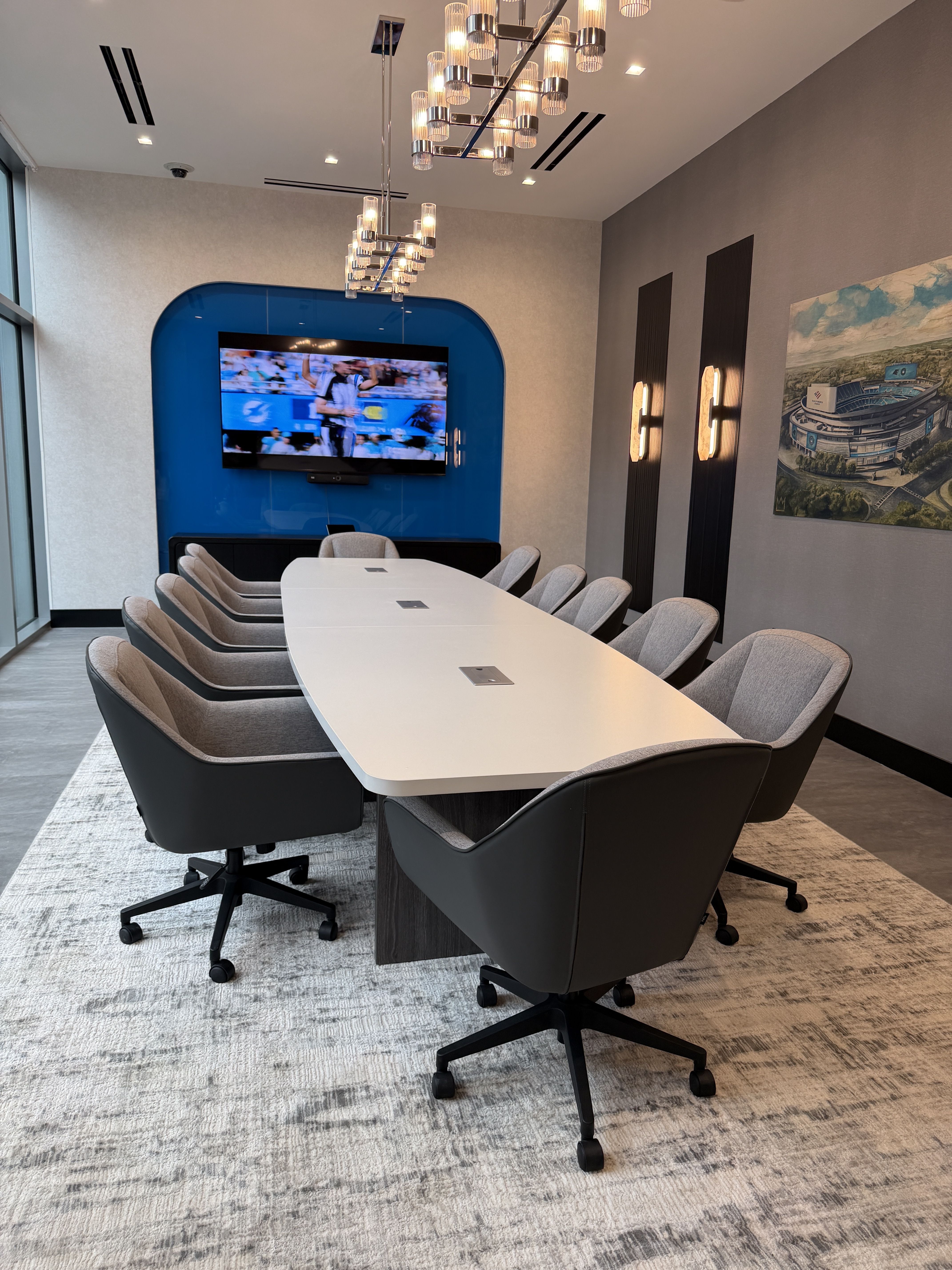 Modern conference room with a long white table and gray upholstered swivel chairs. A wall-mounted TV on a blue curved panel, stylish chandeliers, light gray walls, and a large stadium artwork on the right.