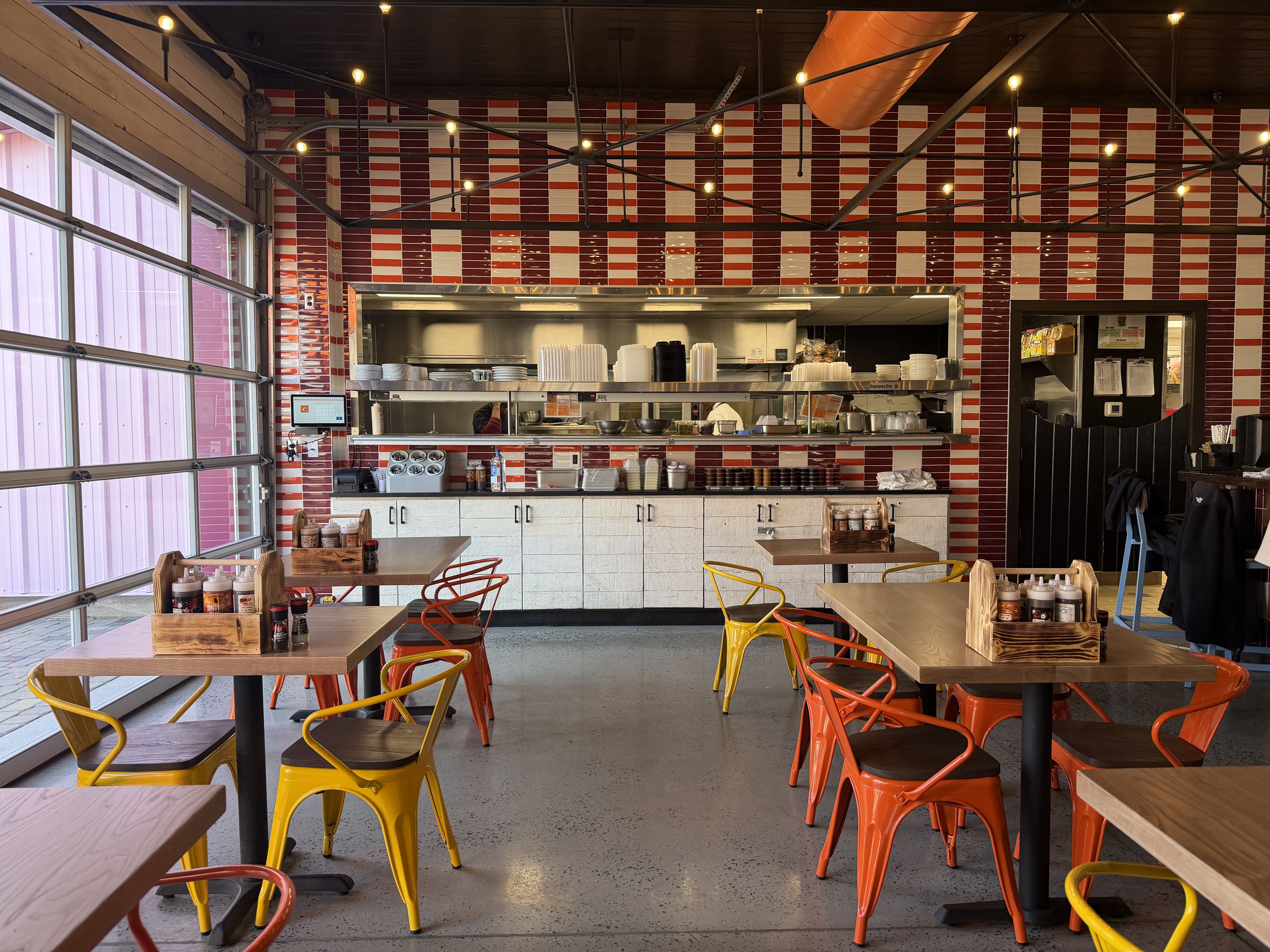 Interior of a restaurant with red and white tiled walls, wooden tables, and yellow and orange metal chairs. Kitchen window and condiment caddies on tables visible.